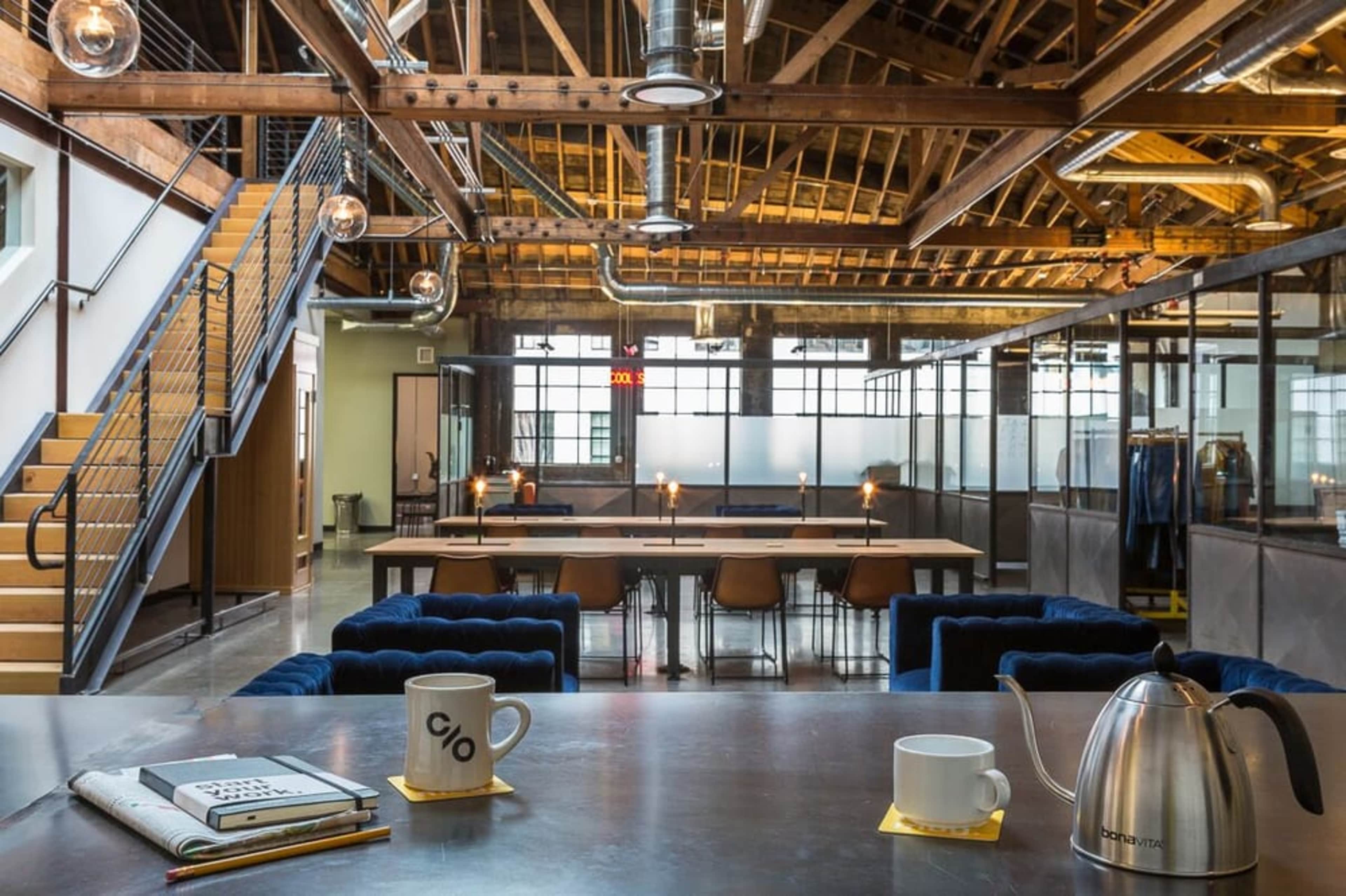 A modern open office space featuring a large communal table, stylish seating, and a wooden ceiling with exposed beams.