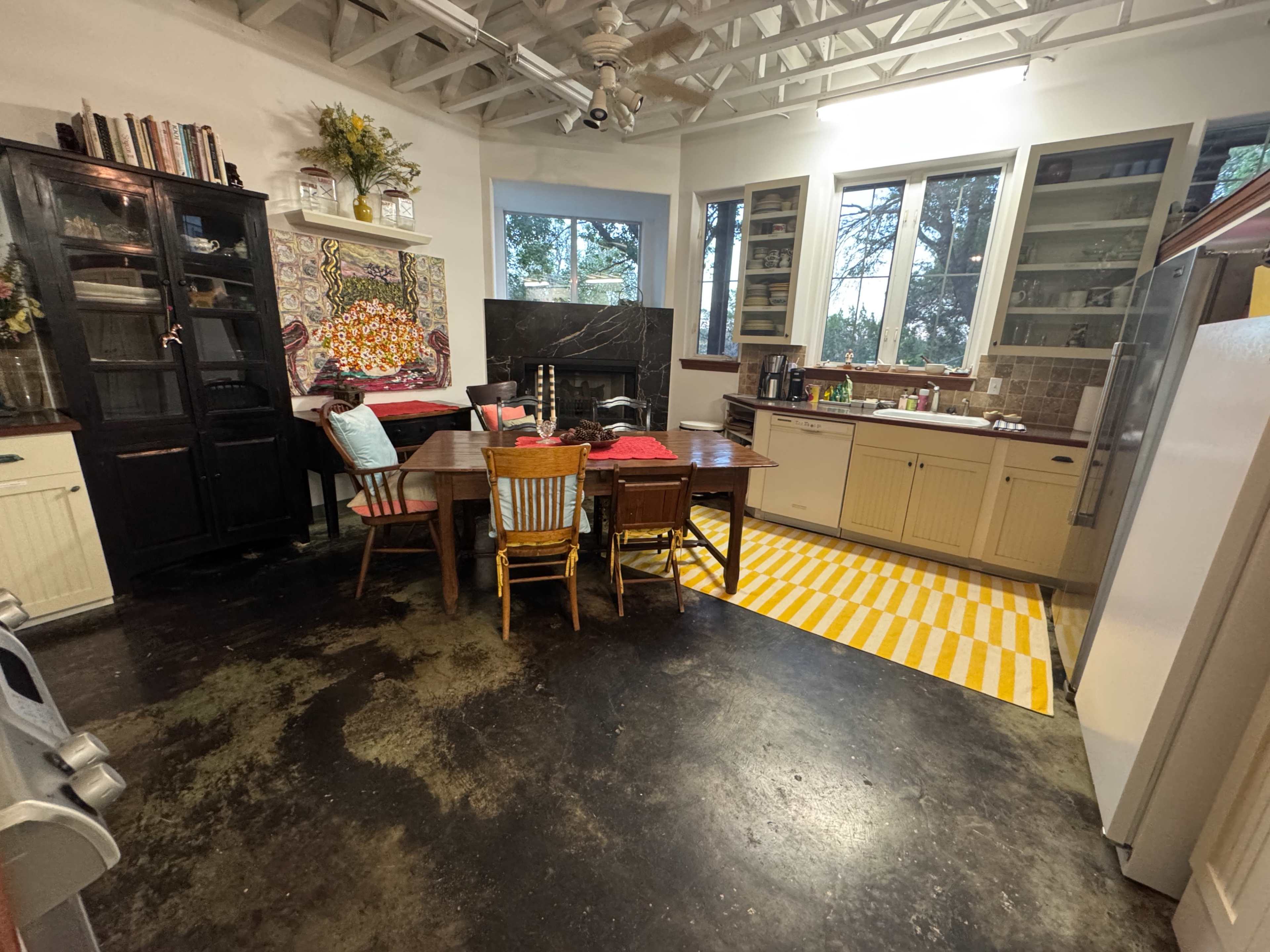 The image shows a kitchen with a wooden dining table surrounded by chairs, featuring a colorful wall art piece and large windows with a view of trees.