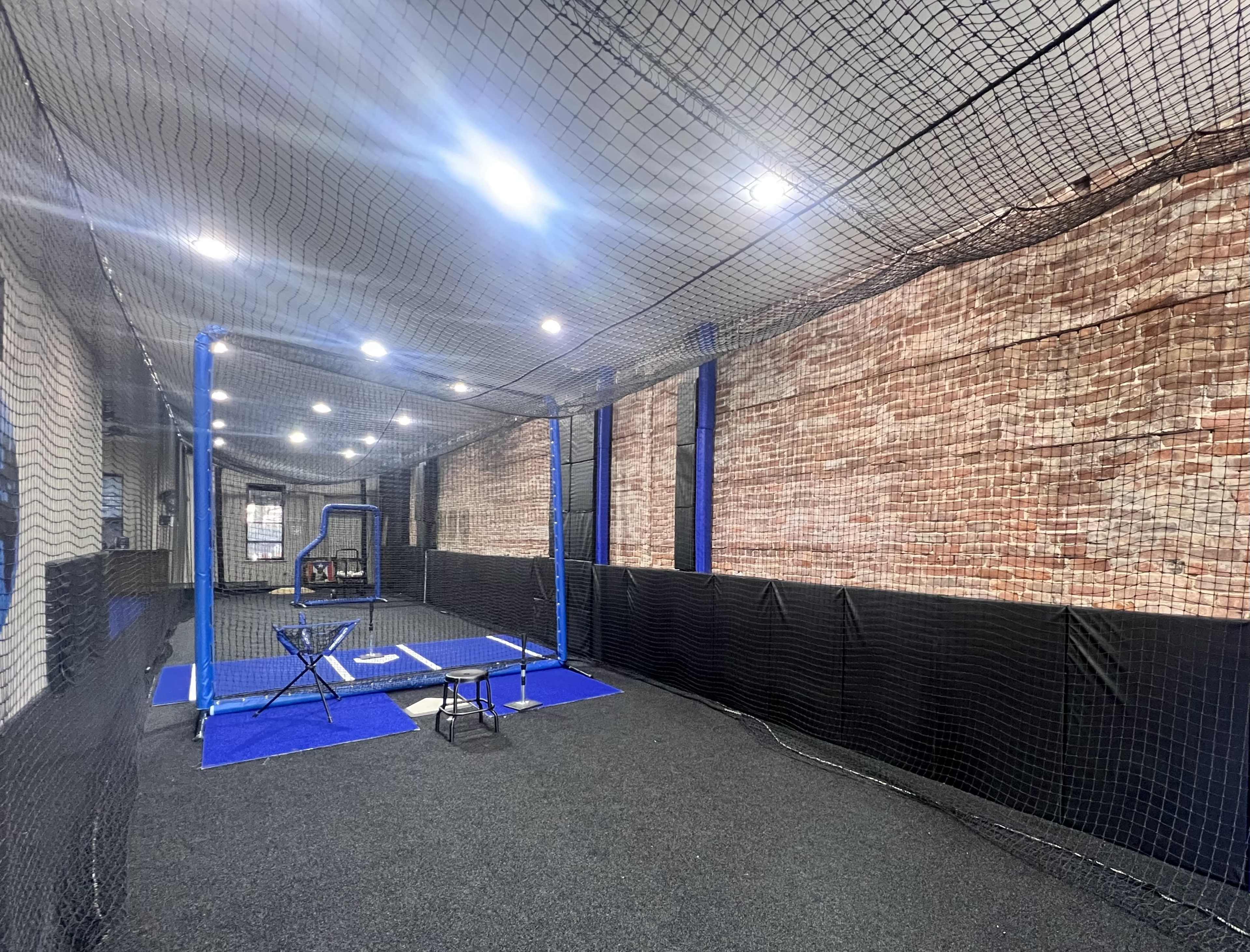 An indoor batting cage facility with a netted area, featuring a batting practice setup and a brick wall backdrop.