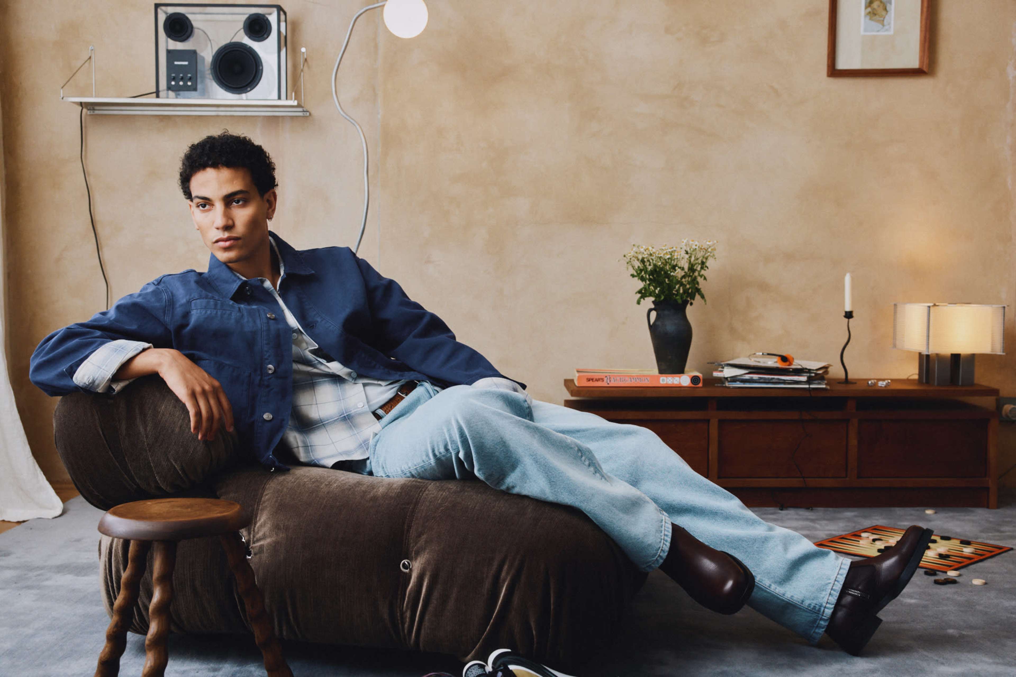 A young man in casual attire lounges on a large, brown bean bag chair in a warmly lit, stylish room with minimal decor, featuring a record player and books on a wooden table.