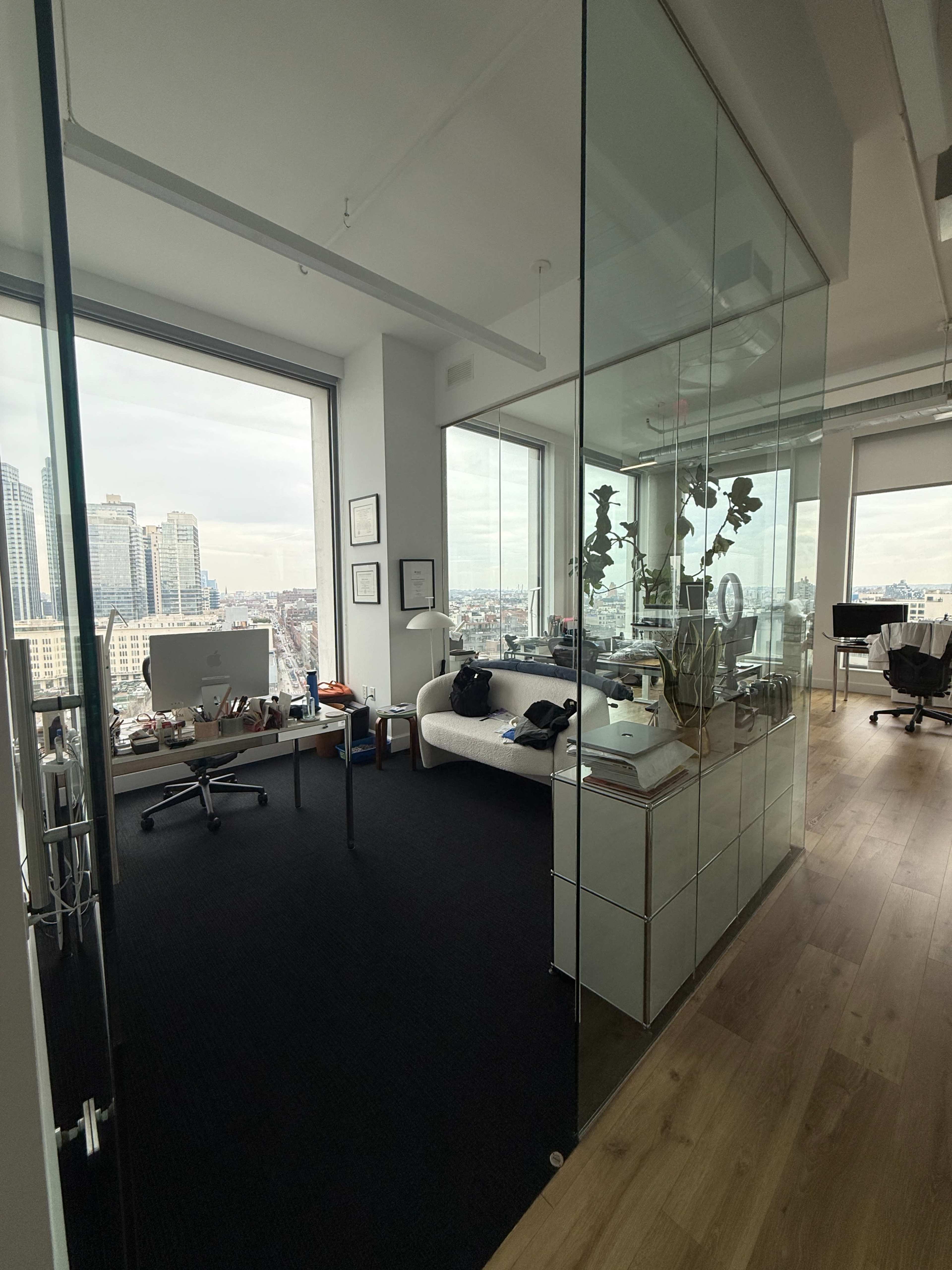The image shows a modern office space with glass walls, featuring a desk, a couch, and a view of a city skyline through large windows.