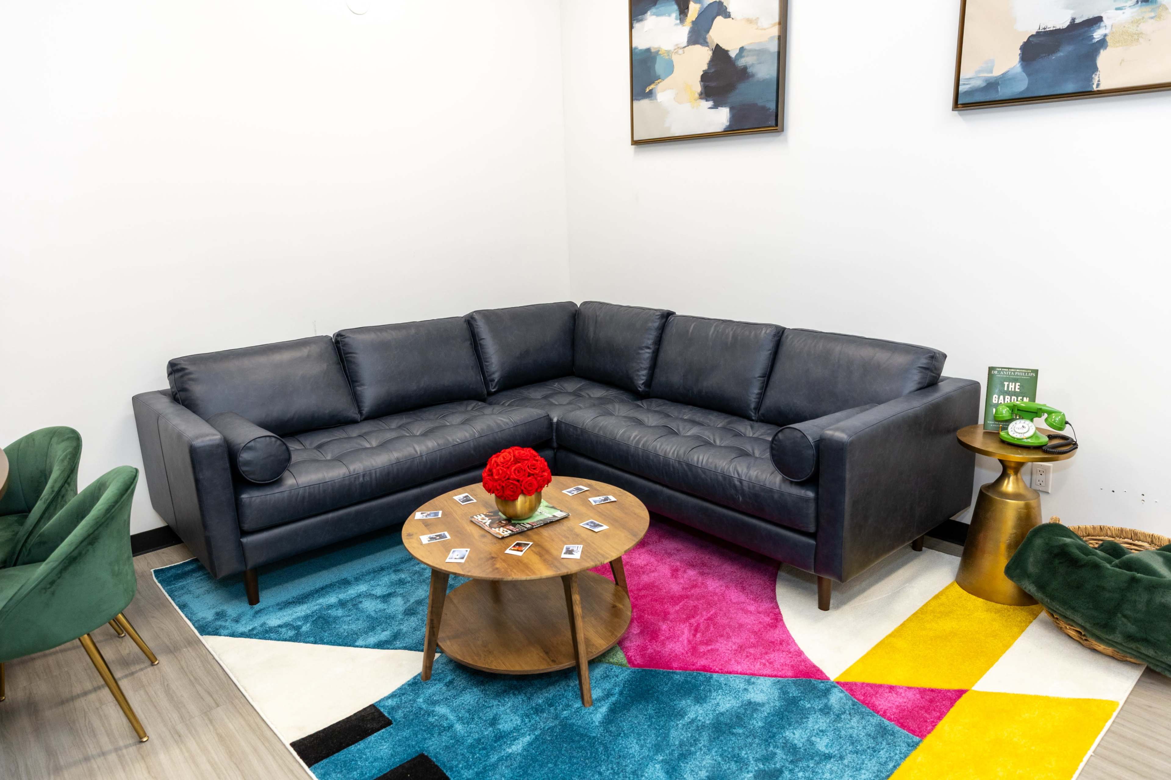 The image shows a modern living room setup featuring a dark blue sectional sofa, a round wooden coffee table adorned with coasters, and a colorful geometric rug.