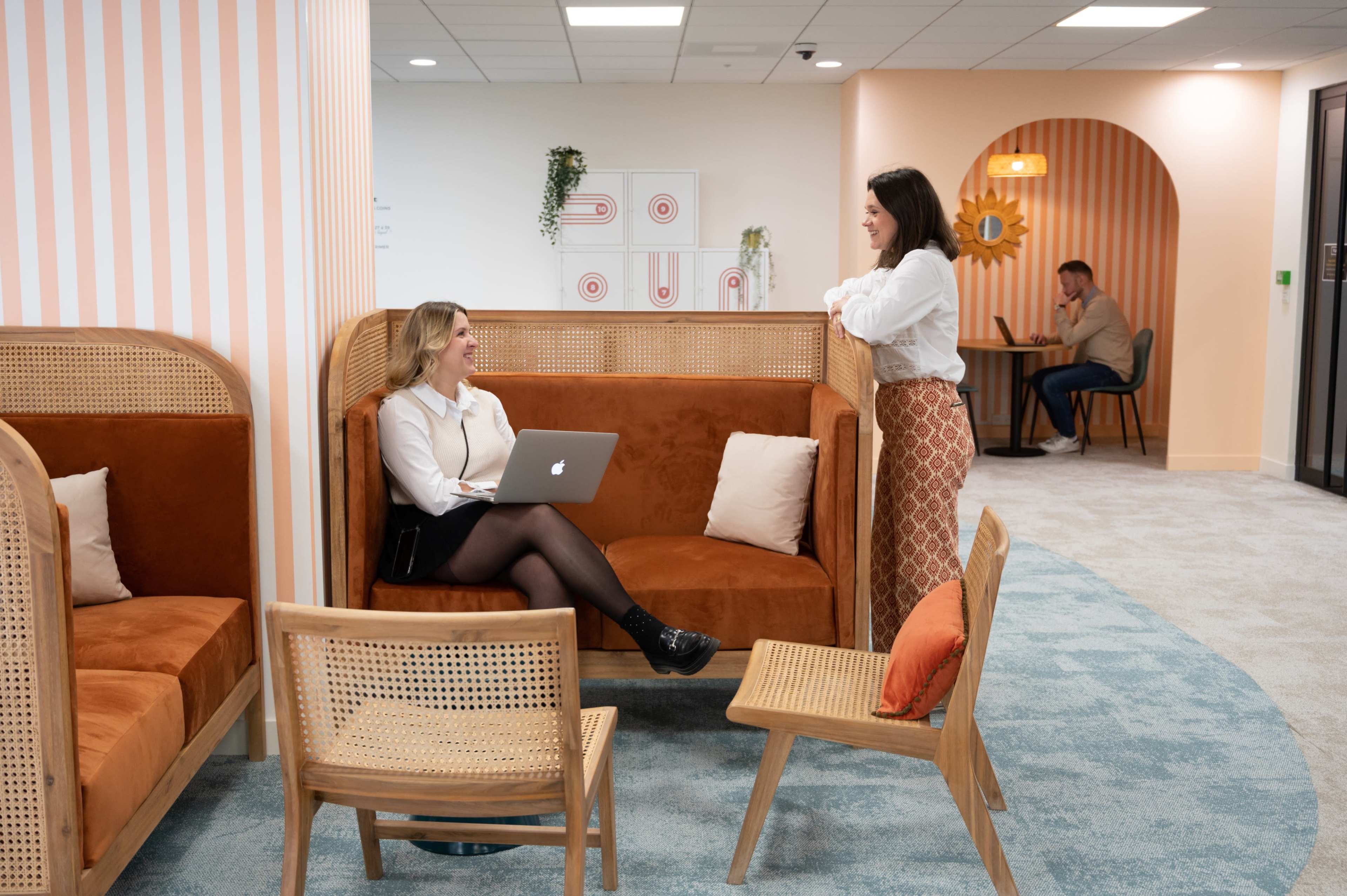 Two women are engaged in conversation in a modern workspace while a man is seated in the background, working at a table.