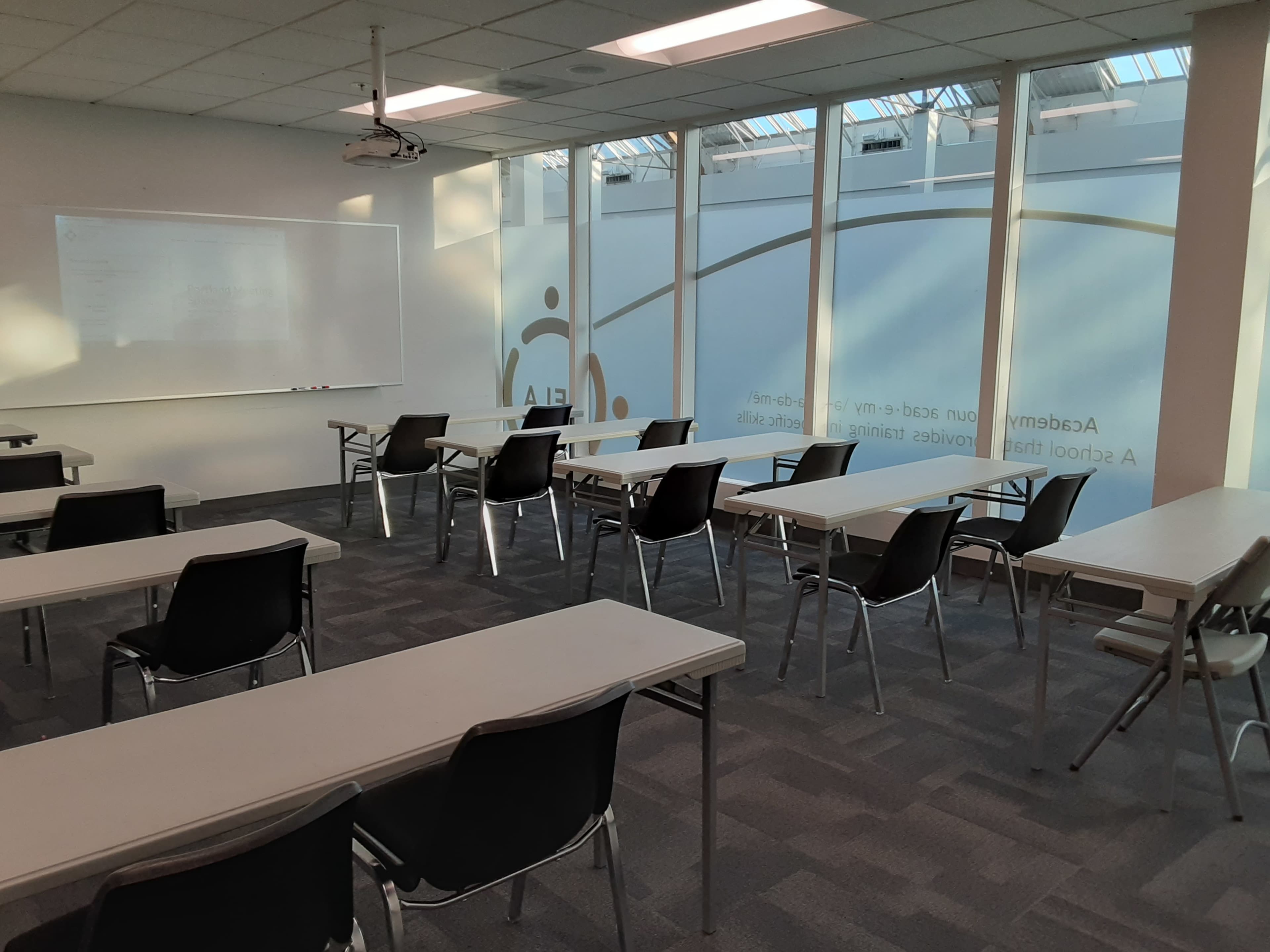 A classroom features several rows of tables and chairs, with large windows letting in natural light.
