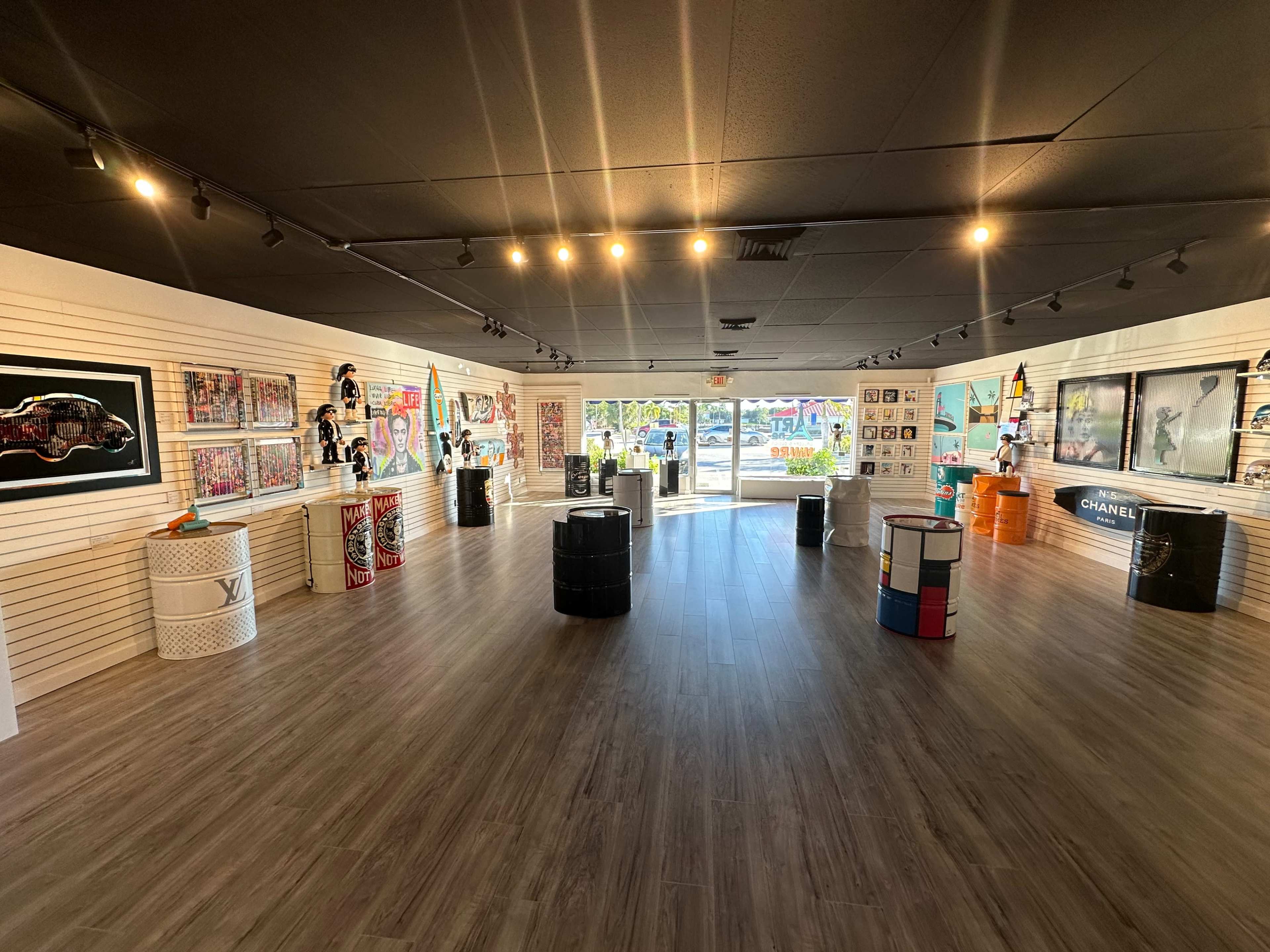 A gallery space featuring various art pieces displayed on walls and several painted barrels arranged in the center.