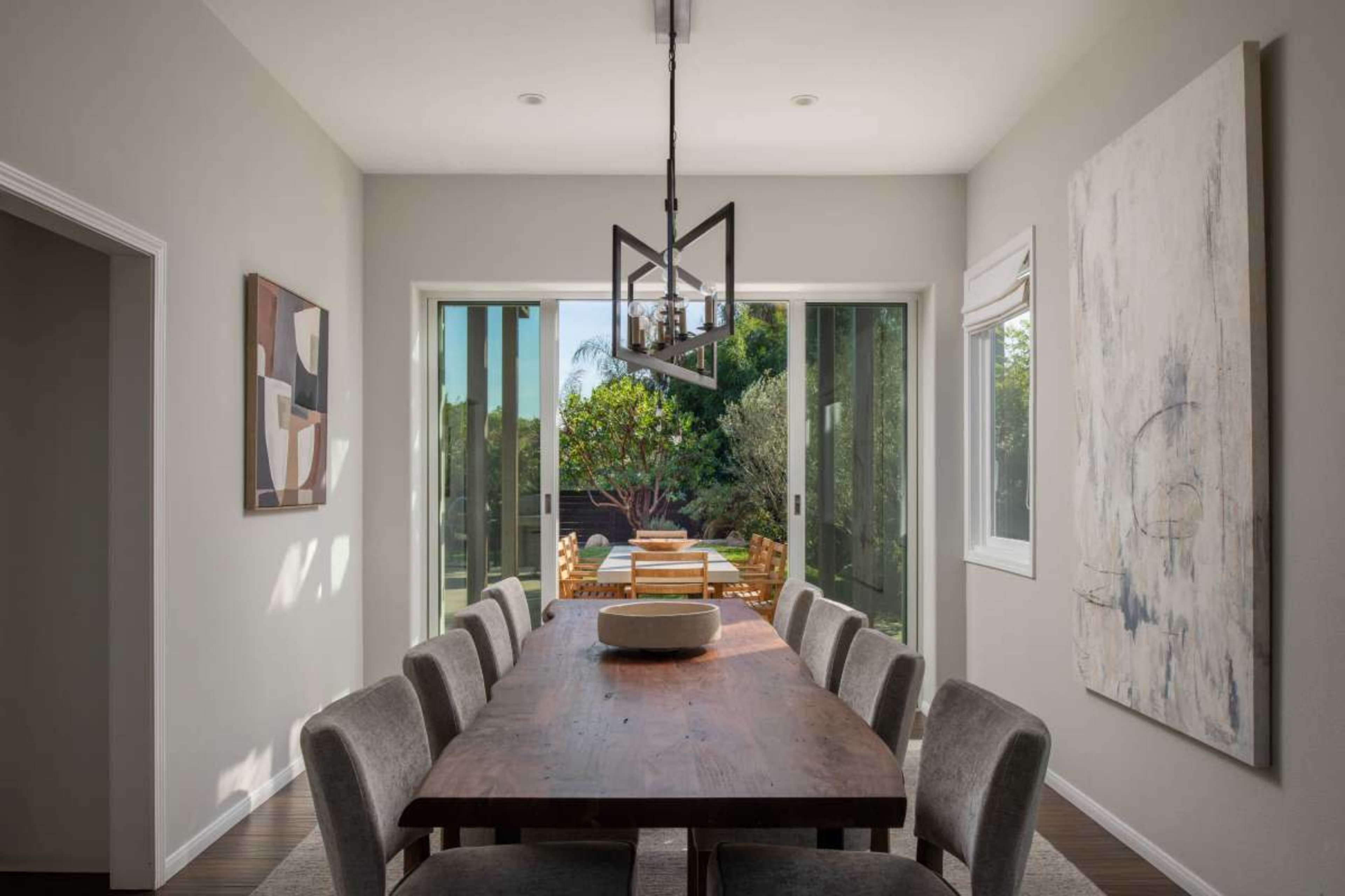 A modern dining room features a long wooden table surrounded by upholstered chairs, with large windows that open to a lush outdoor area.