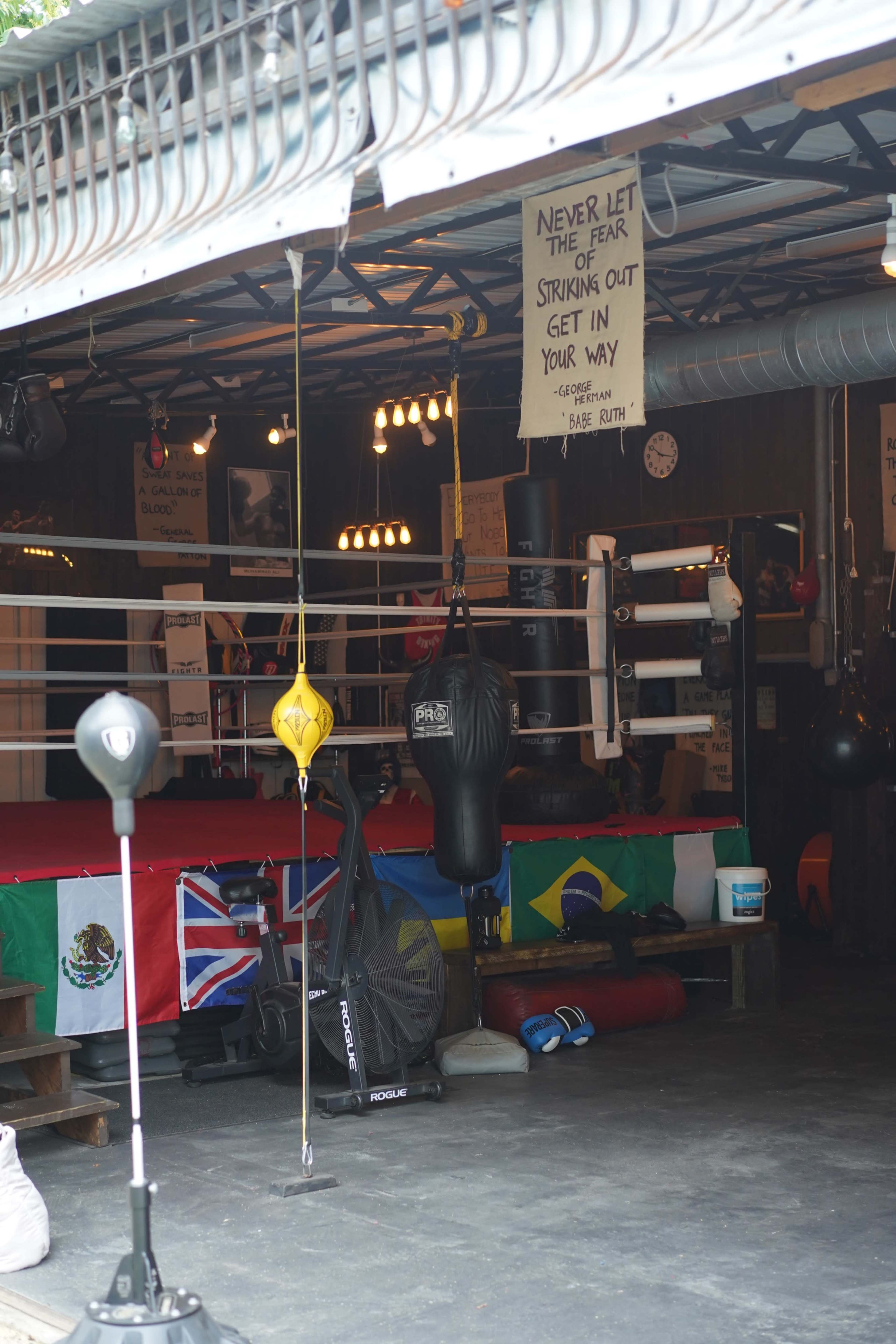 A boxing gym features a ring surrounded by flags from various countries, heavy bags hanging, and motivational quotes on the walls.