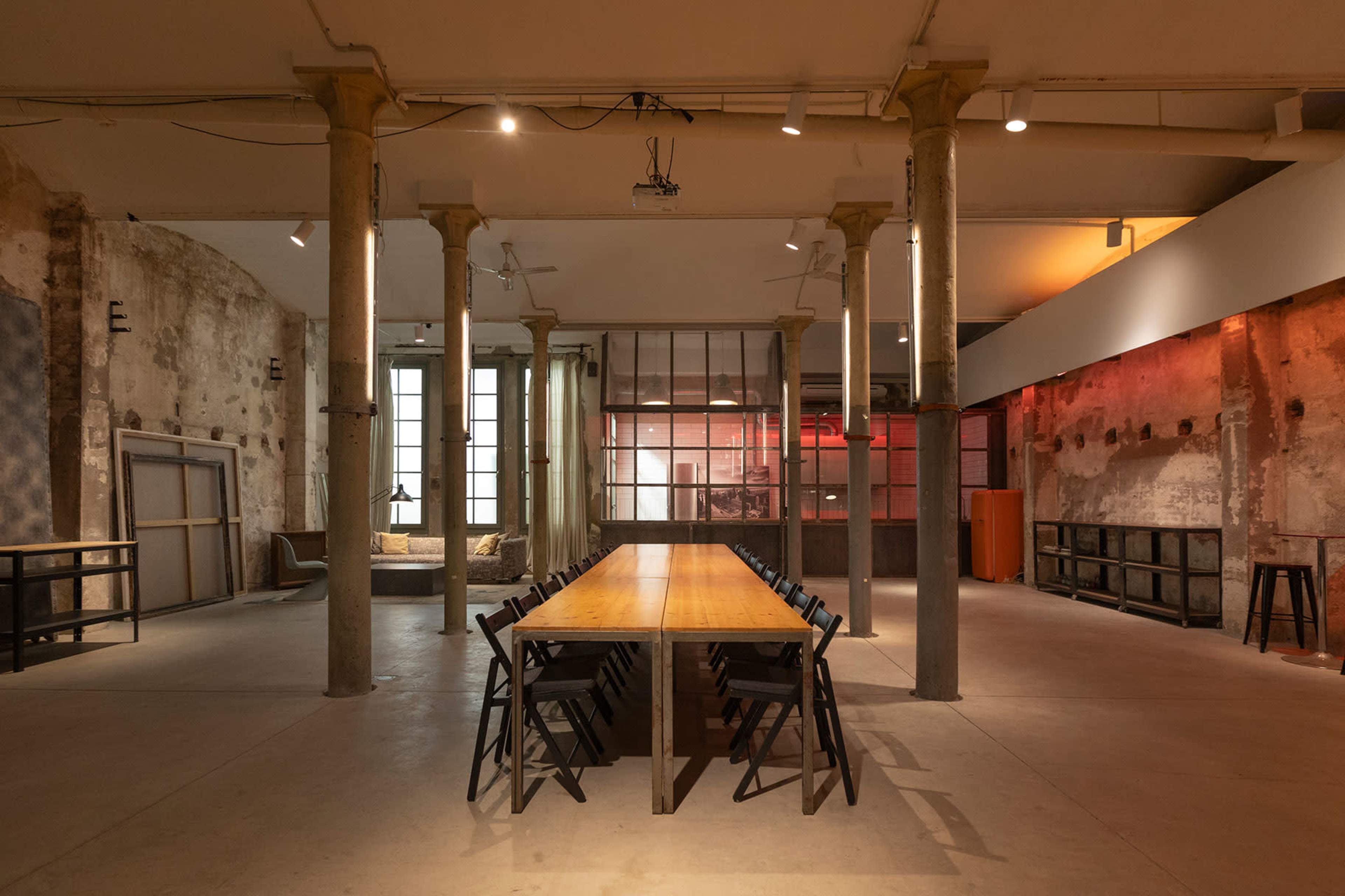 The image shows an empty industrial-style room with a long wooden table surrounded by black chairs, tall columns, and large windows.