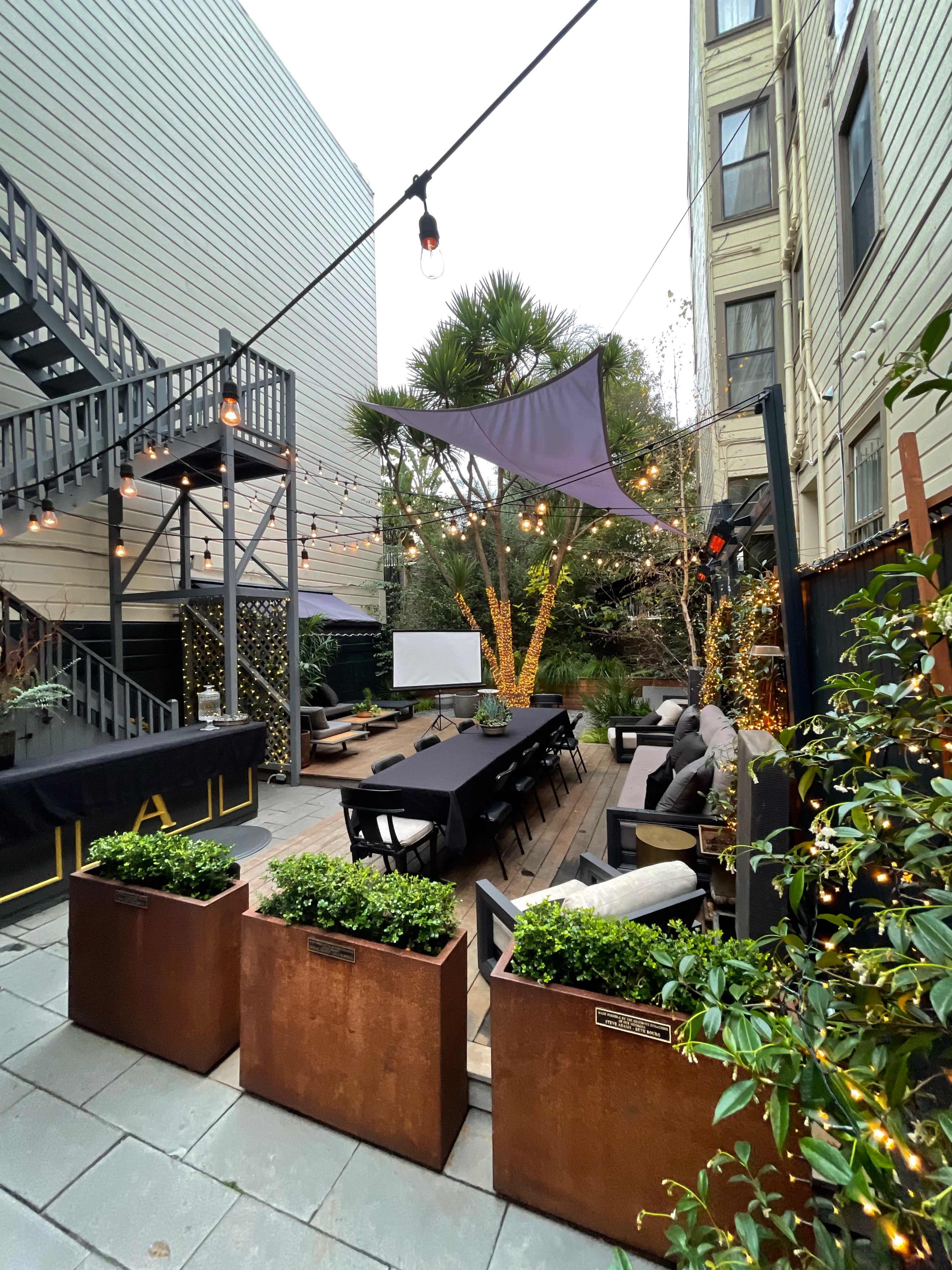 A courtyard with a long dining table surrounded by planters, string lights overhead, and a projector screen at one end.