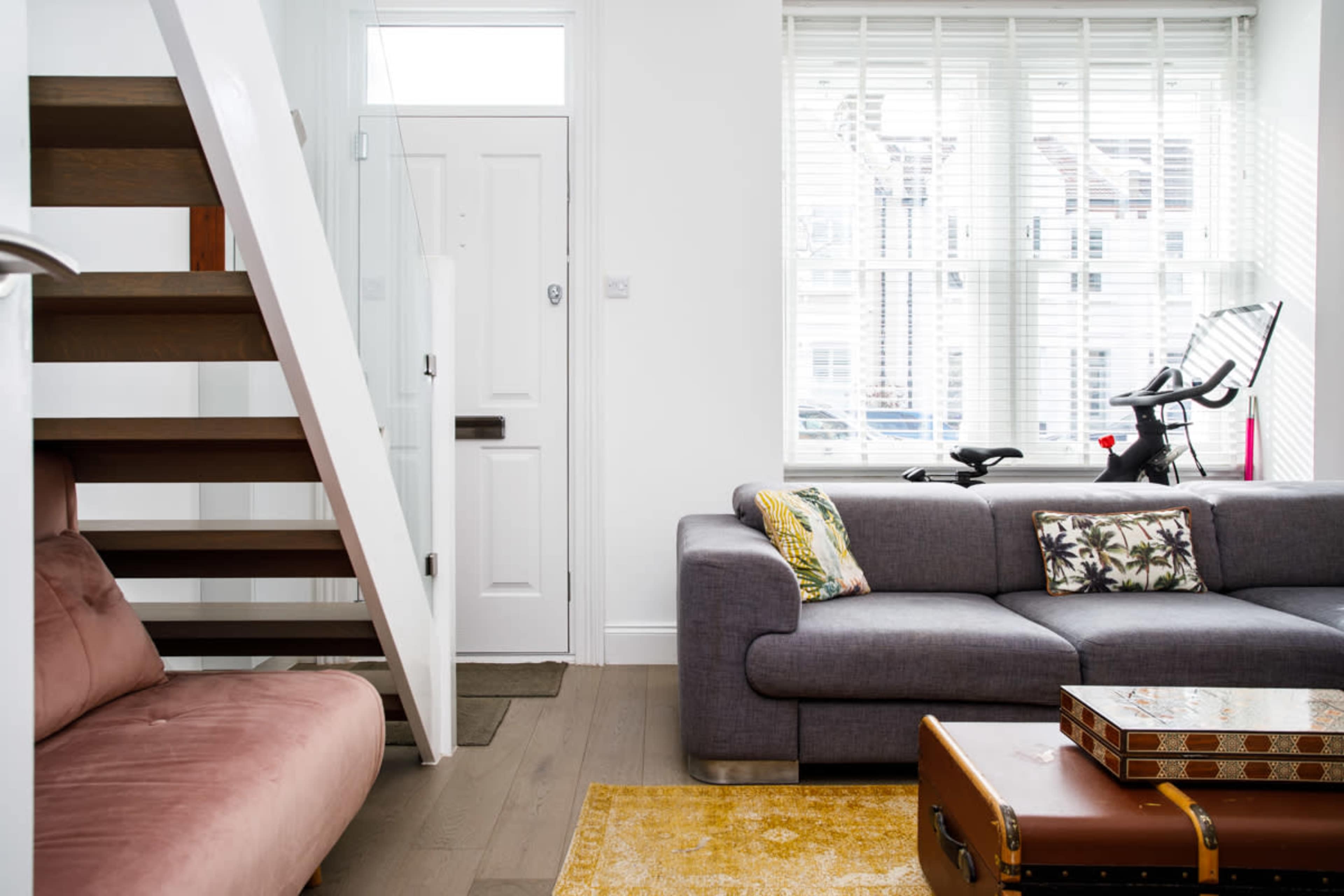 The image depicts a bright living room with a staircase, a gray sofa, a pink chair, and a stationary exercise bike near the window.