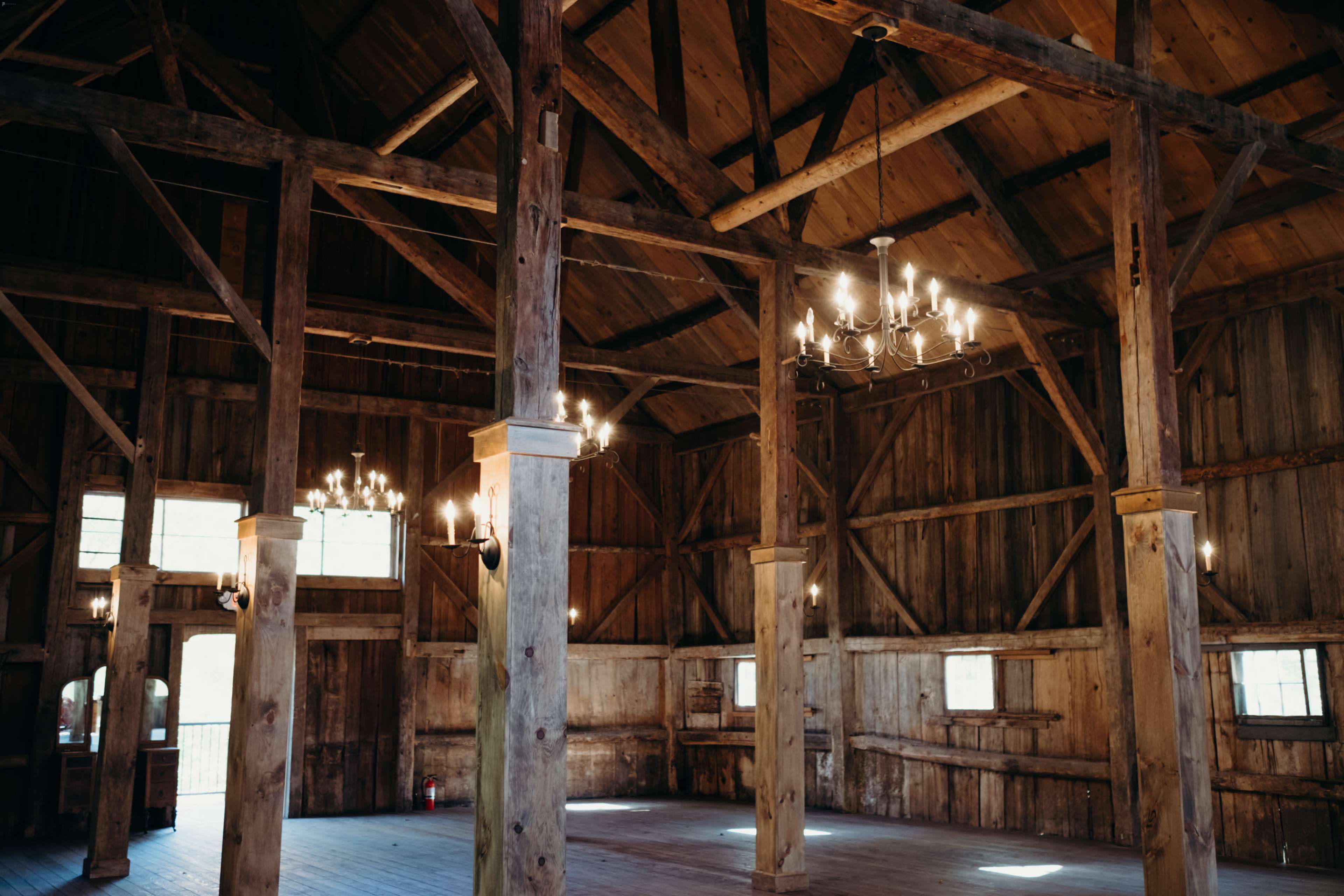 The interior of a rustic barn with wooden beams, chandeliers hanging from the ceiling, and large windows allowing natural light to enter.
