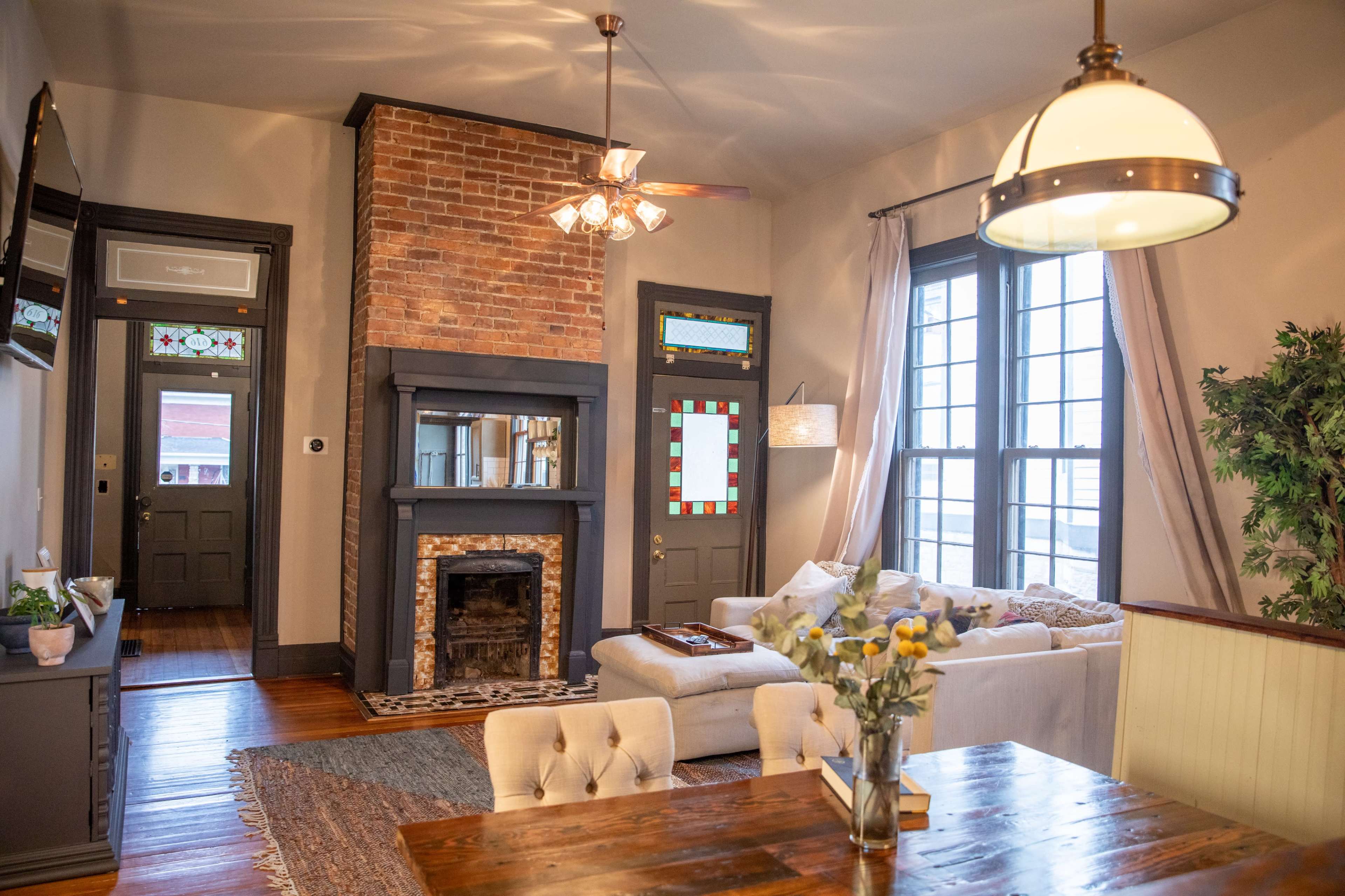 A cozy living room featuring a brick fireplace, light-colored furniture, and large windows with decorative stained glass.