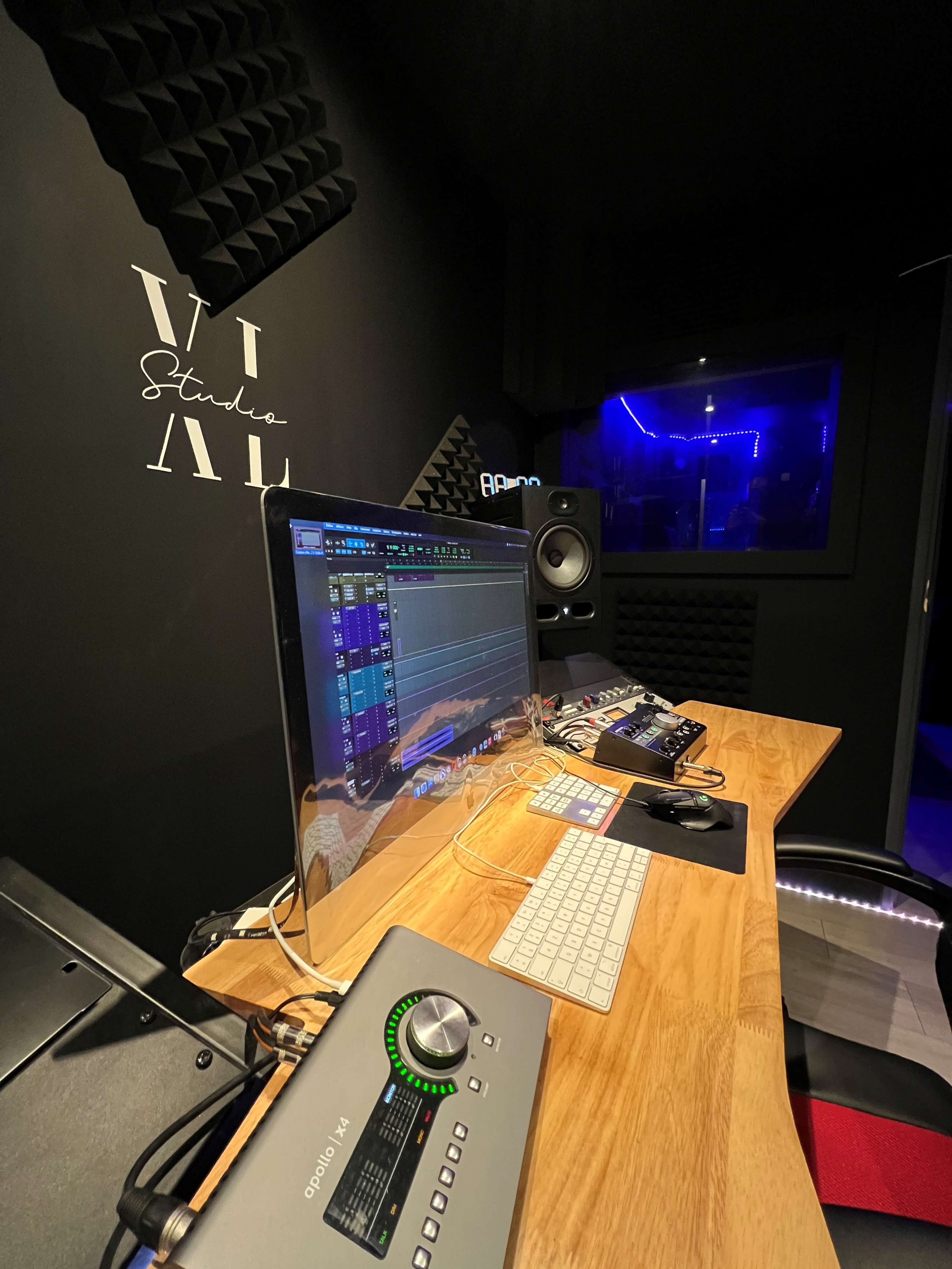 A music production studio featuring a computer monitor with digital audio software, a keyboard, and a sound mixer on a wooden desk against a dark wall.