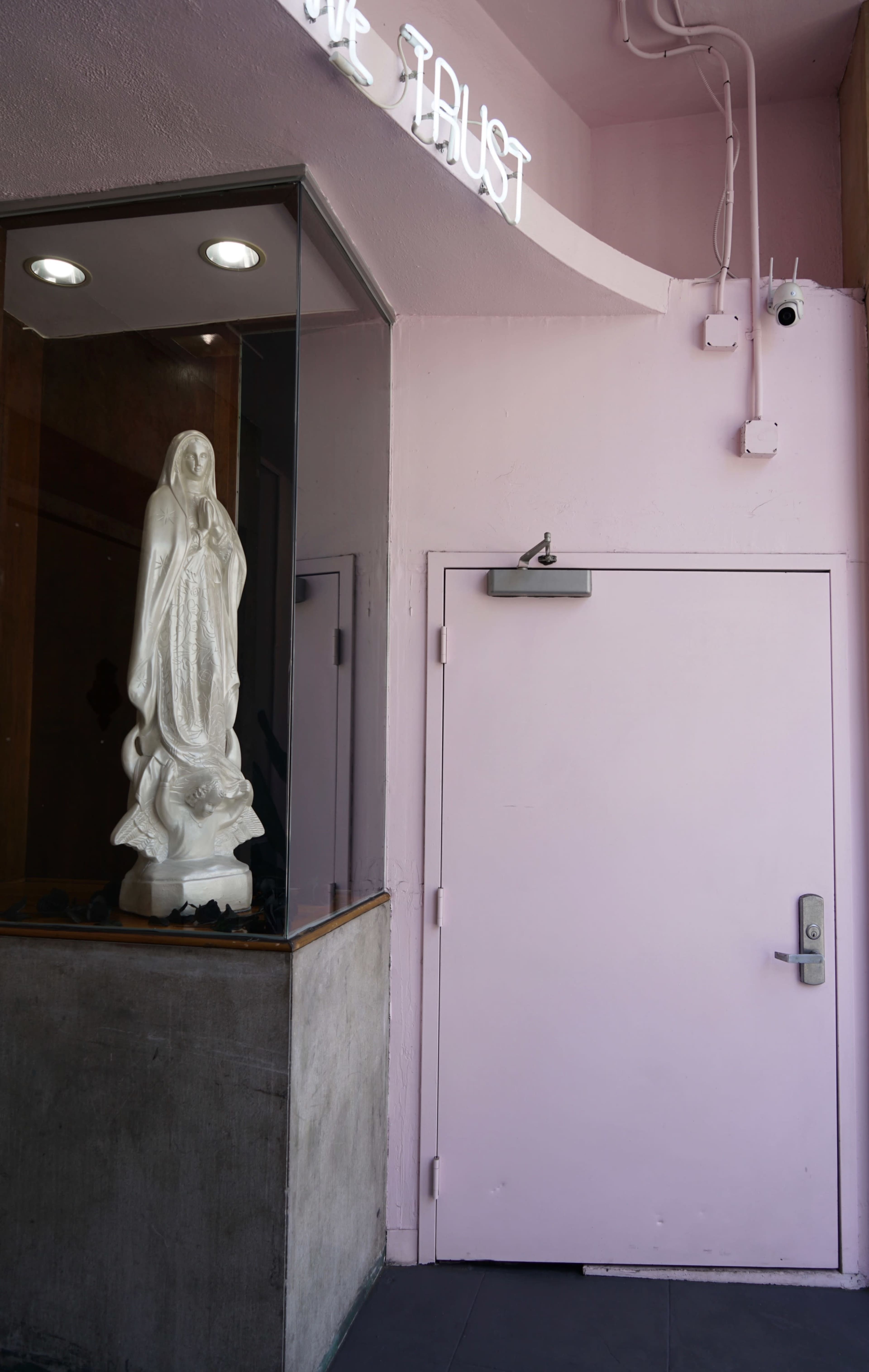 A white statue of a figure stands in a glass enclosure next to a pink door on a wall.
