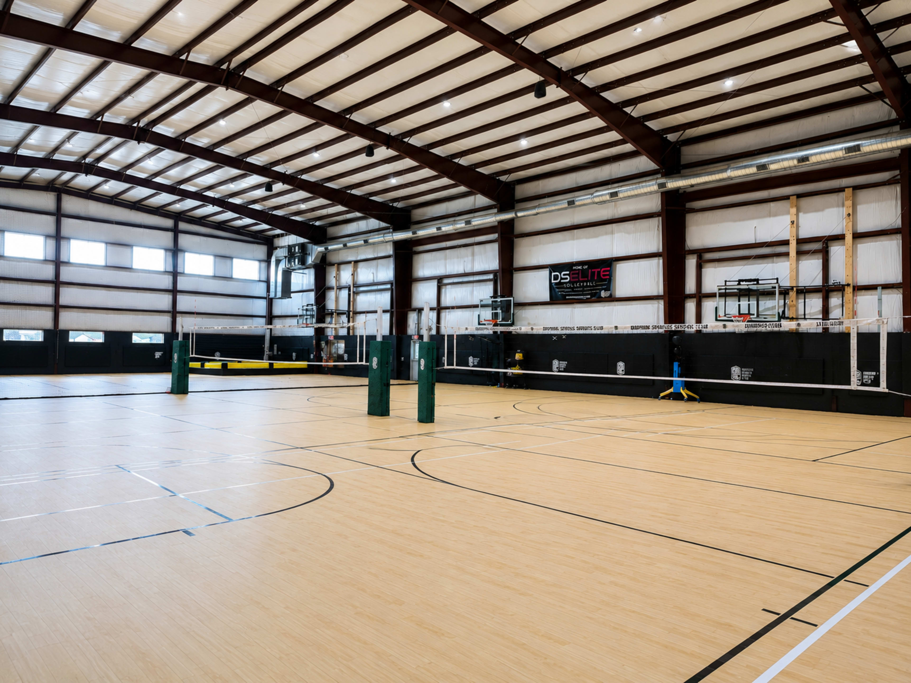 An indoor sports facility features a hardwood floor with multiple volleyball courts, basketball hoops, and large windows letting in natural light.