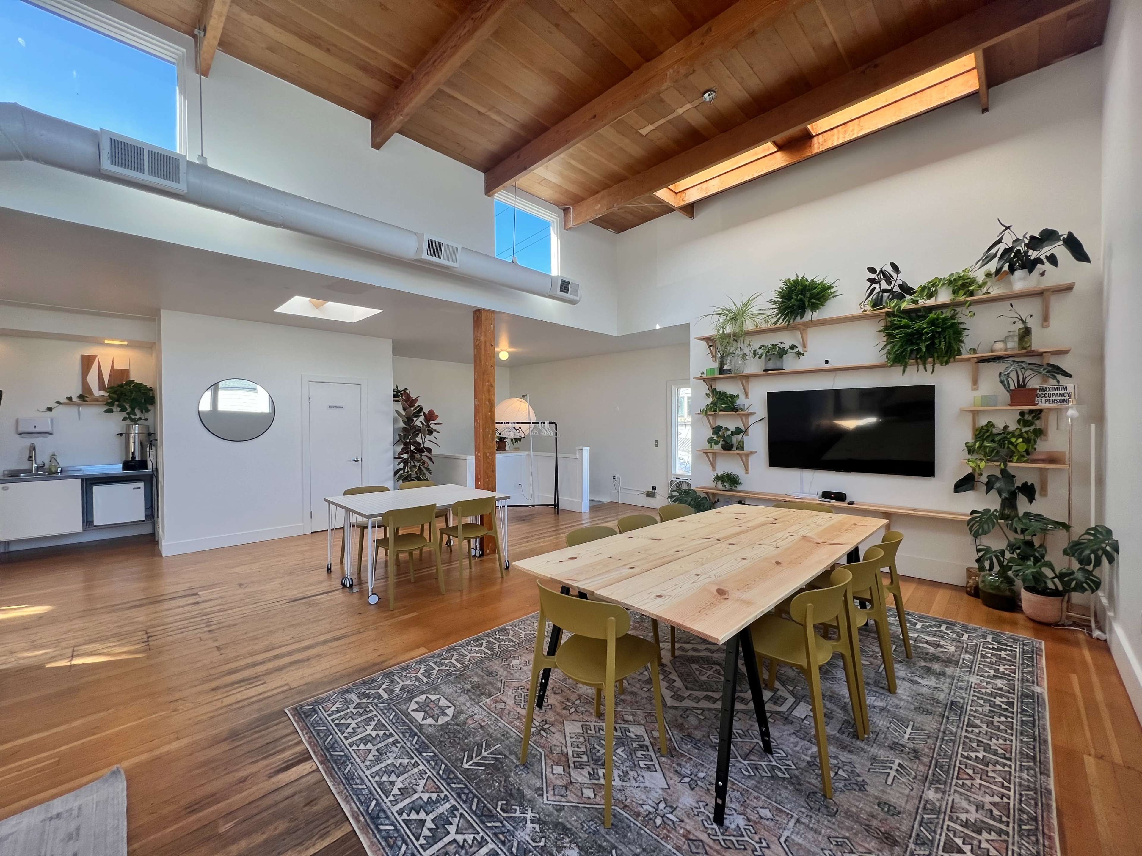 A spacious room features a dining table surrounded by chairs, a television mounted on a wall with plants, and a kitchenette in the background.