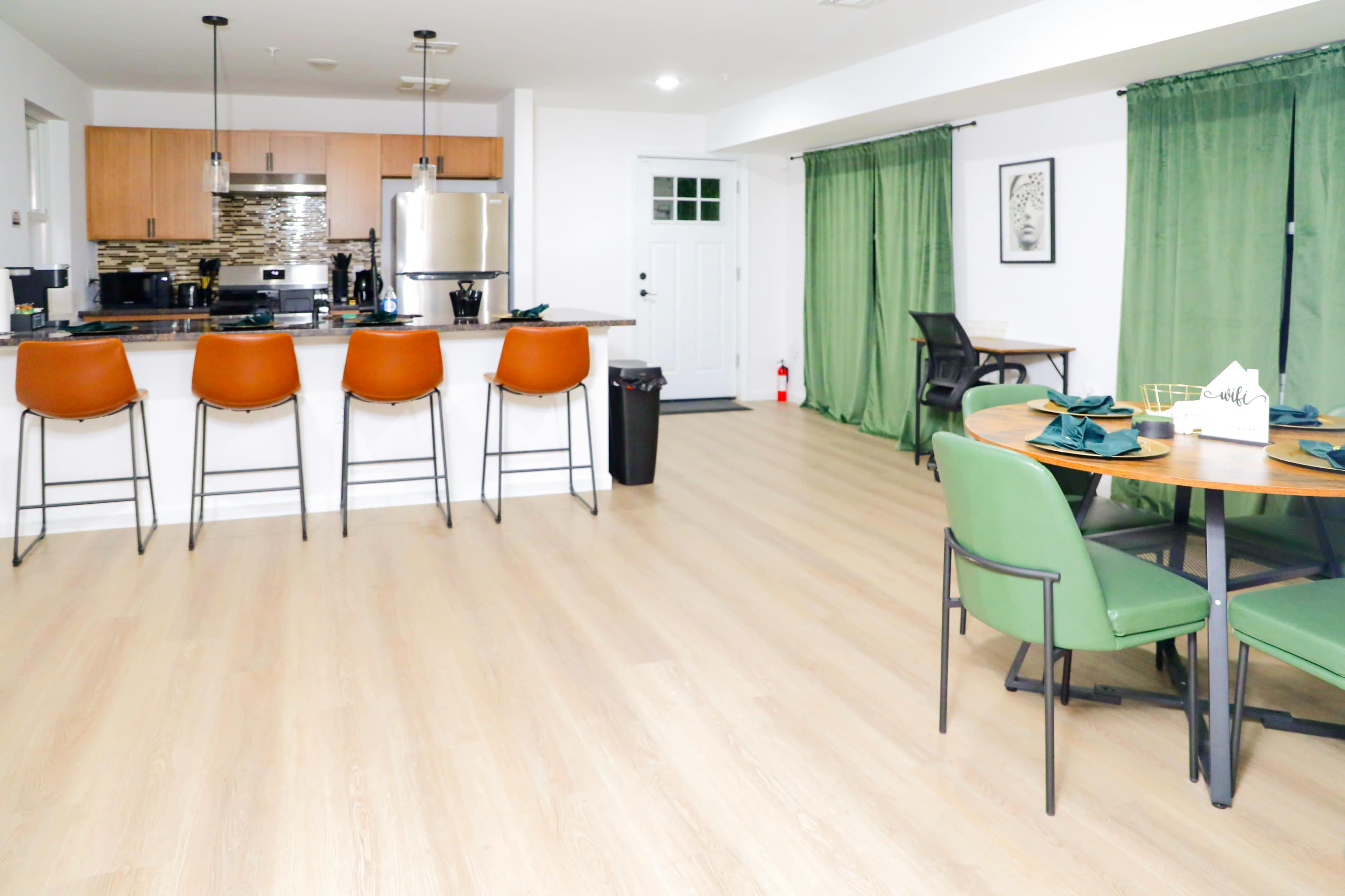 The image shows a modern interior featuring a kitchen with bar stools, a dining area with green chairs, and large windows covered by green curtains.