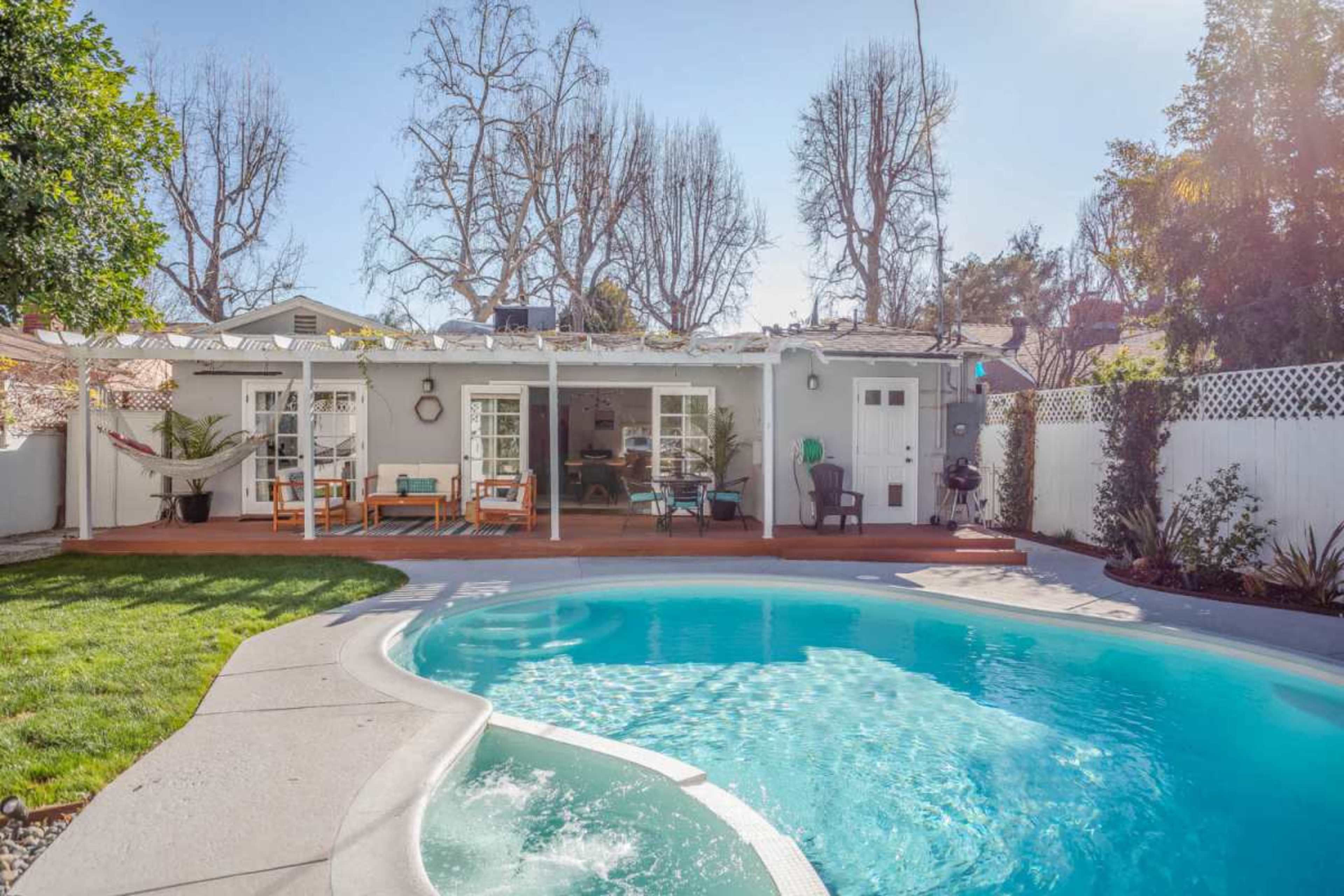 The image shows a backyard with a swimming pool, green grass, and a house with a patio furnished with chairs and a hammock, surrounded by trees.