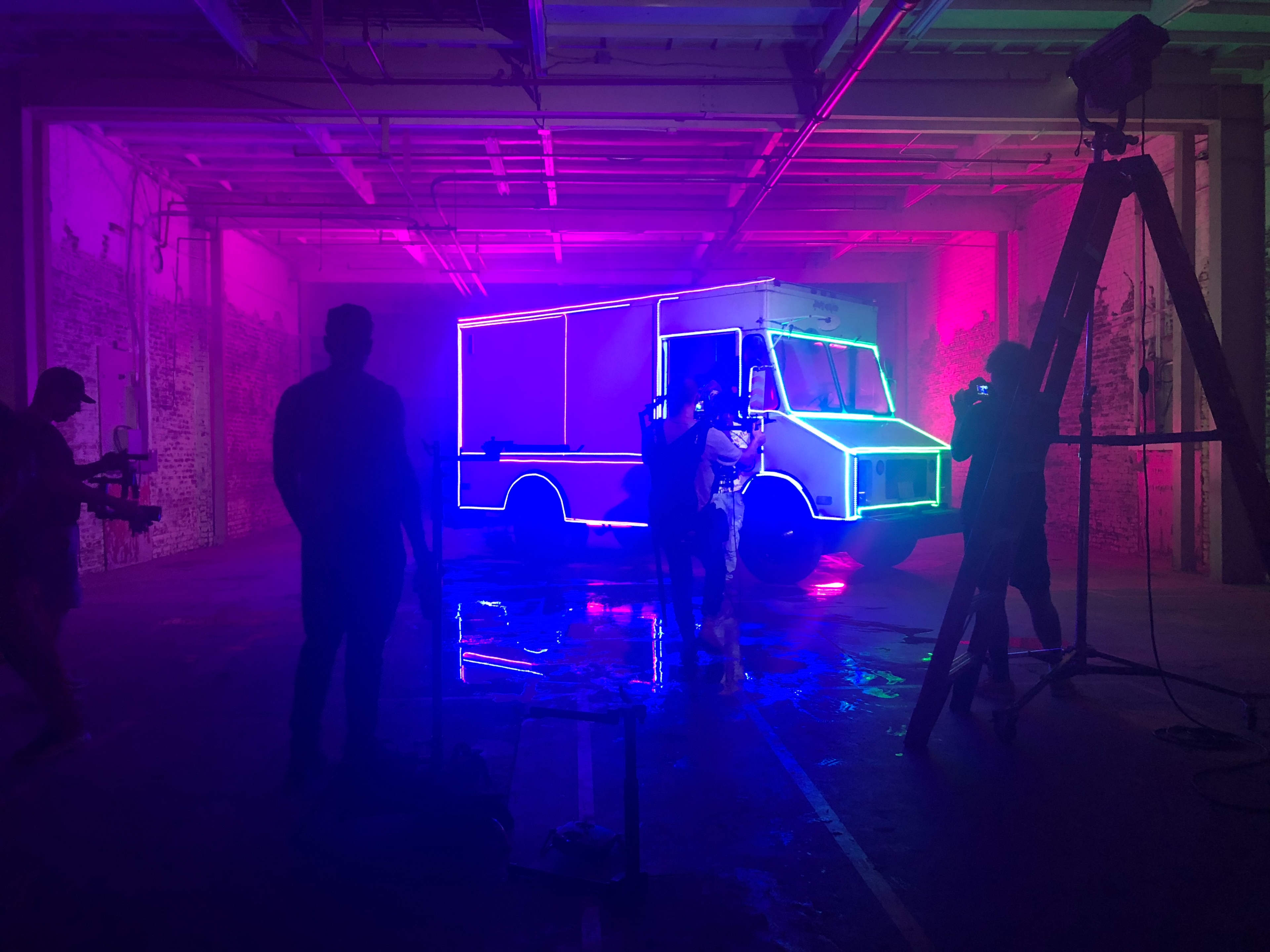 A brightly lit truck with neon outlines is parked in a dimly lit room, while crew members set up equipment for a shoot.