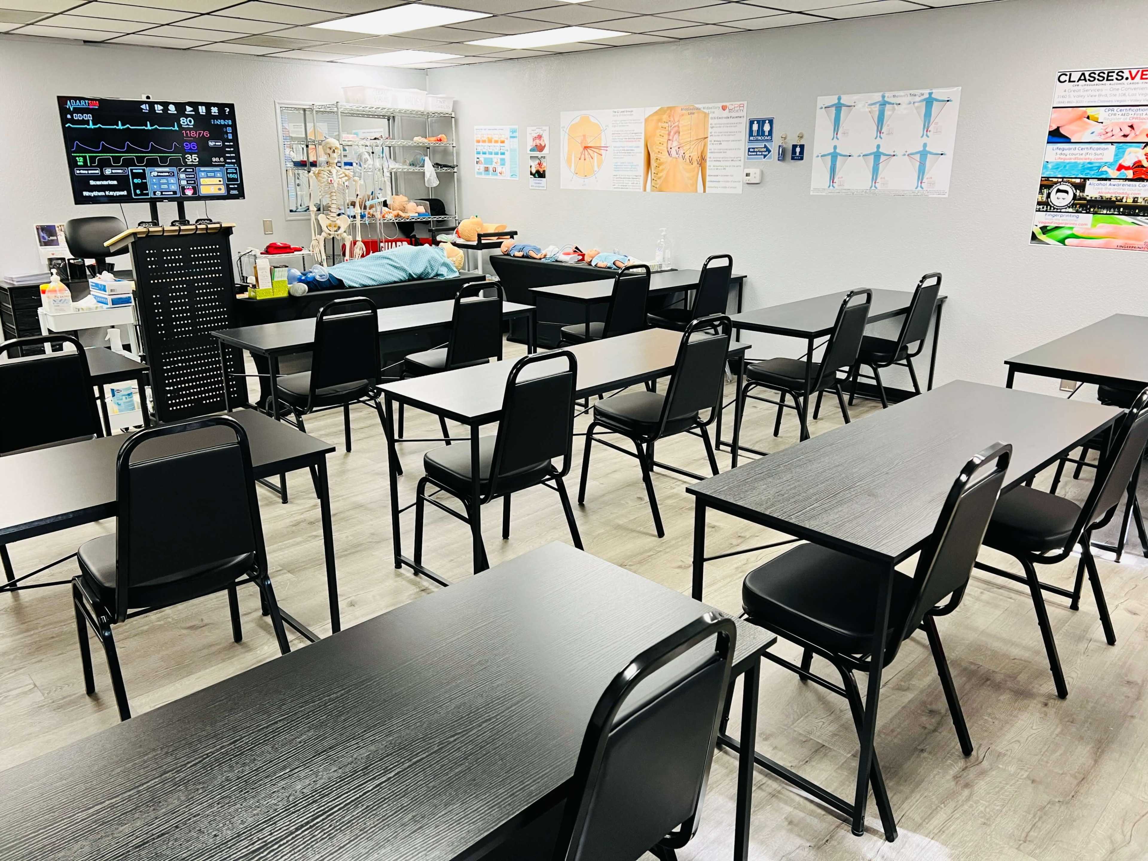 The image shows a classroom setup with black tables and chairs arranged in rows, featuring educational posters and medical training equipment on the walls.