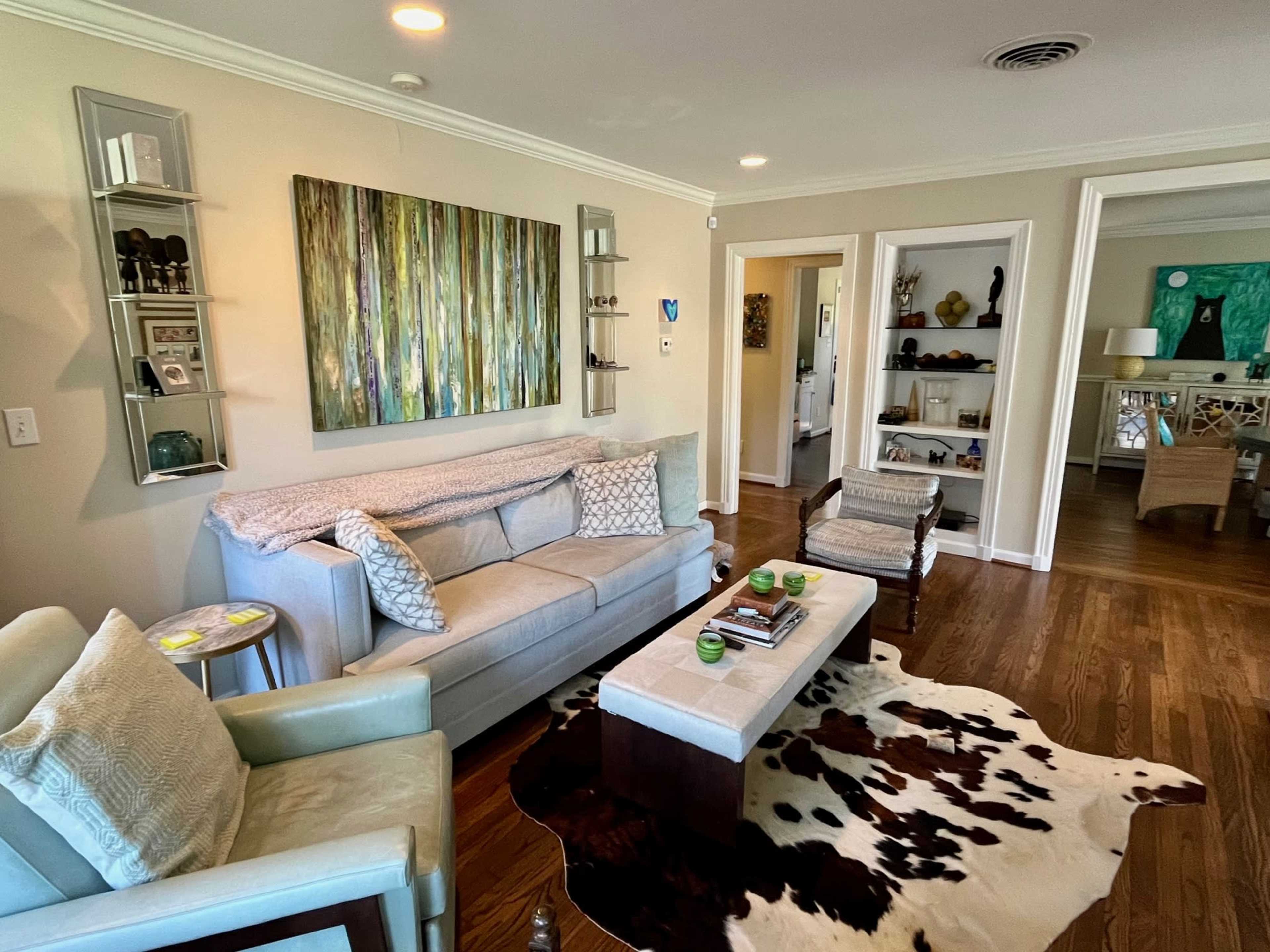 A living room with a light-colored sofa, a wooden coffee table on a cowhide rug, and a green abstract painting on the wall, with various decorative items displayed on shelves.