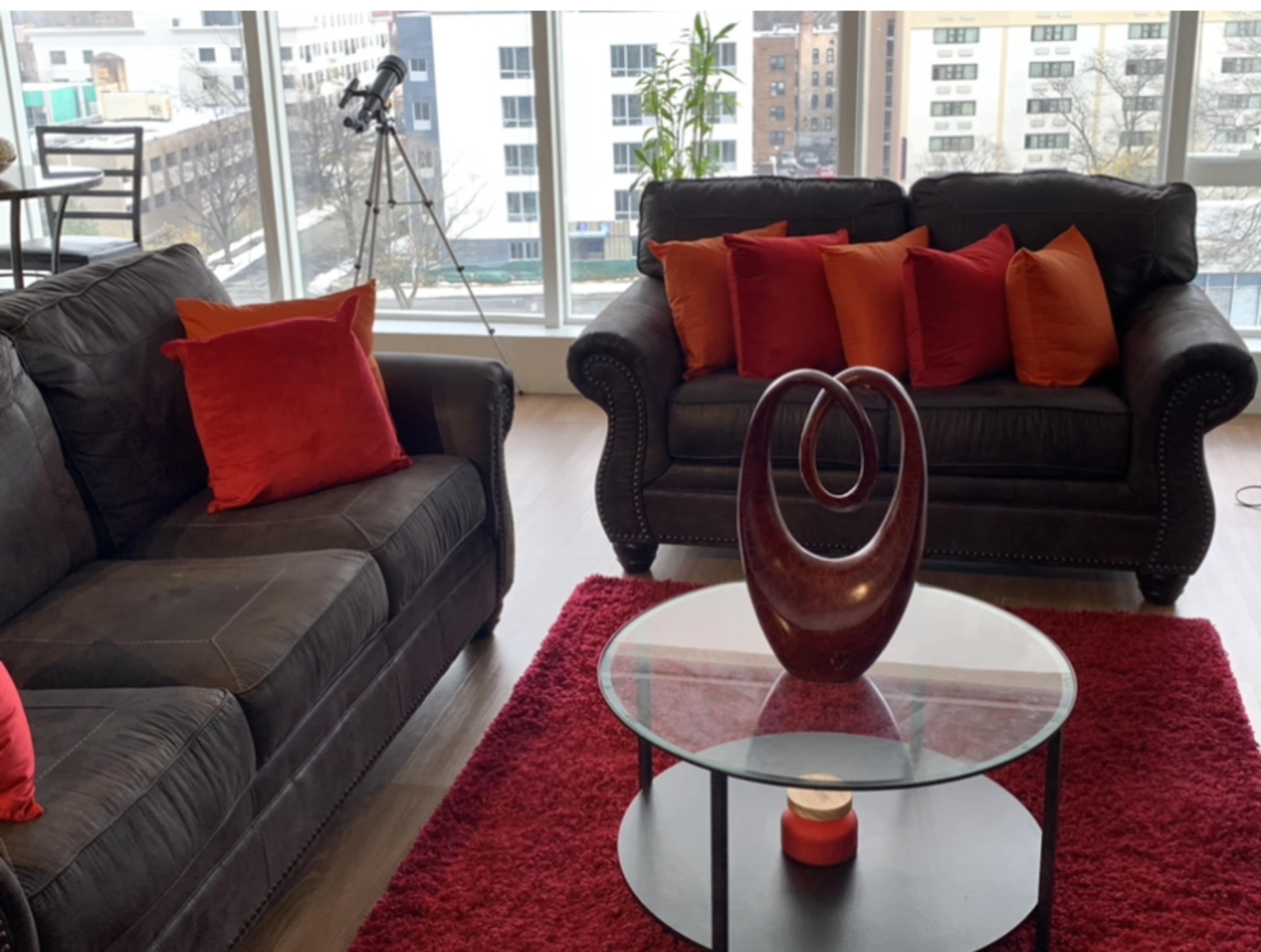 A modern living room features two dark sofas adorned with red and orange pillows, a glass coffee table with a decorative vase, and large windows displaying an urban view.