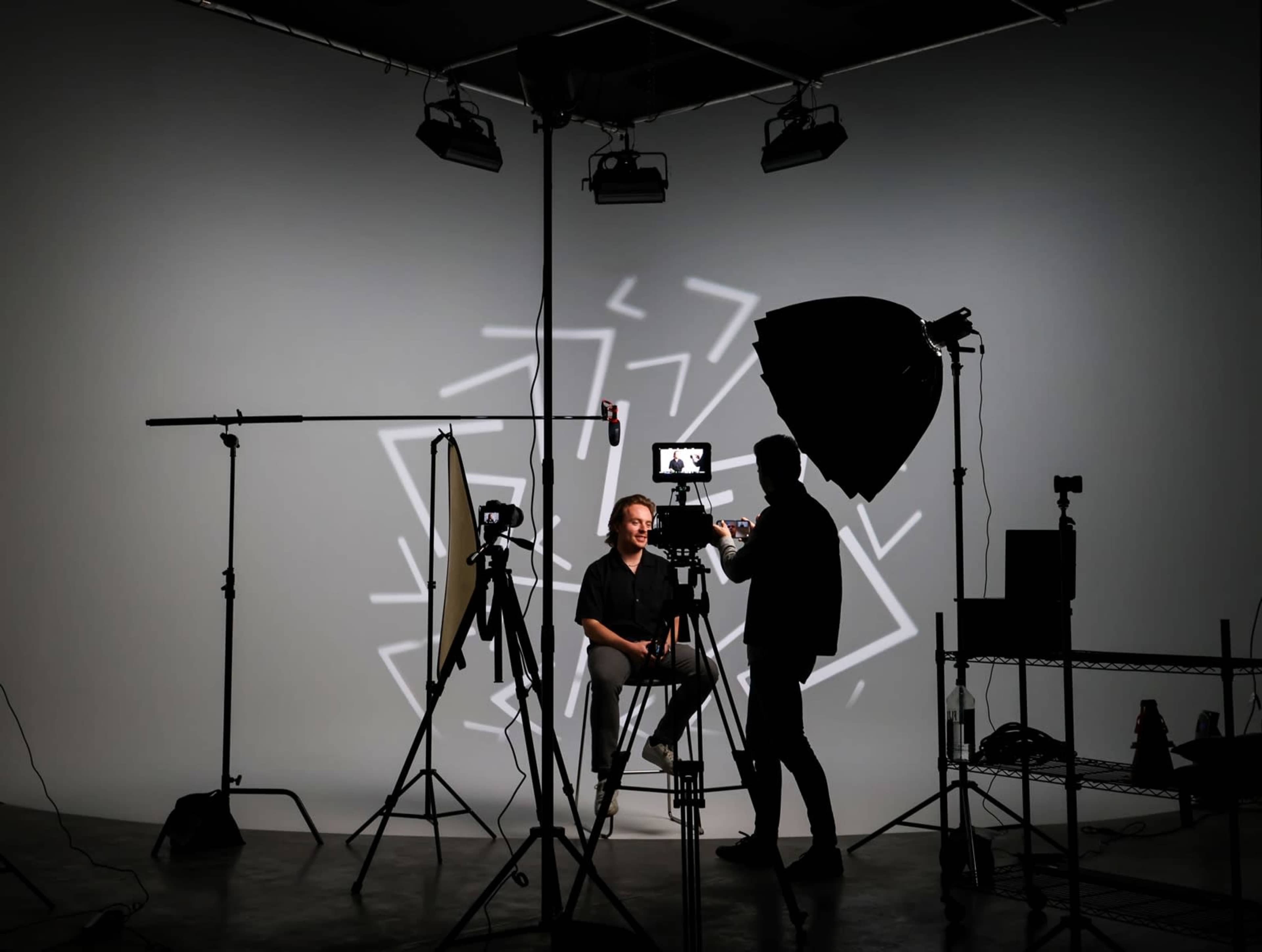 A person sits on a stool in a photography studio surrounded by lighting equipment and camera setups against a backdrop with geometric patterns.