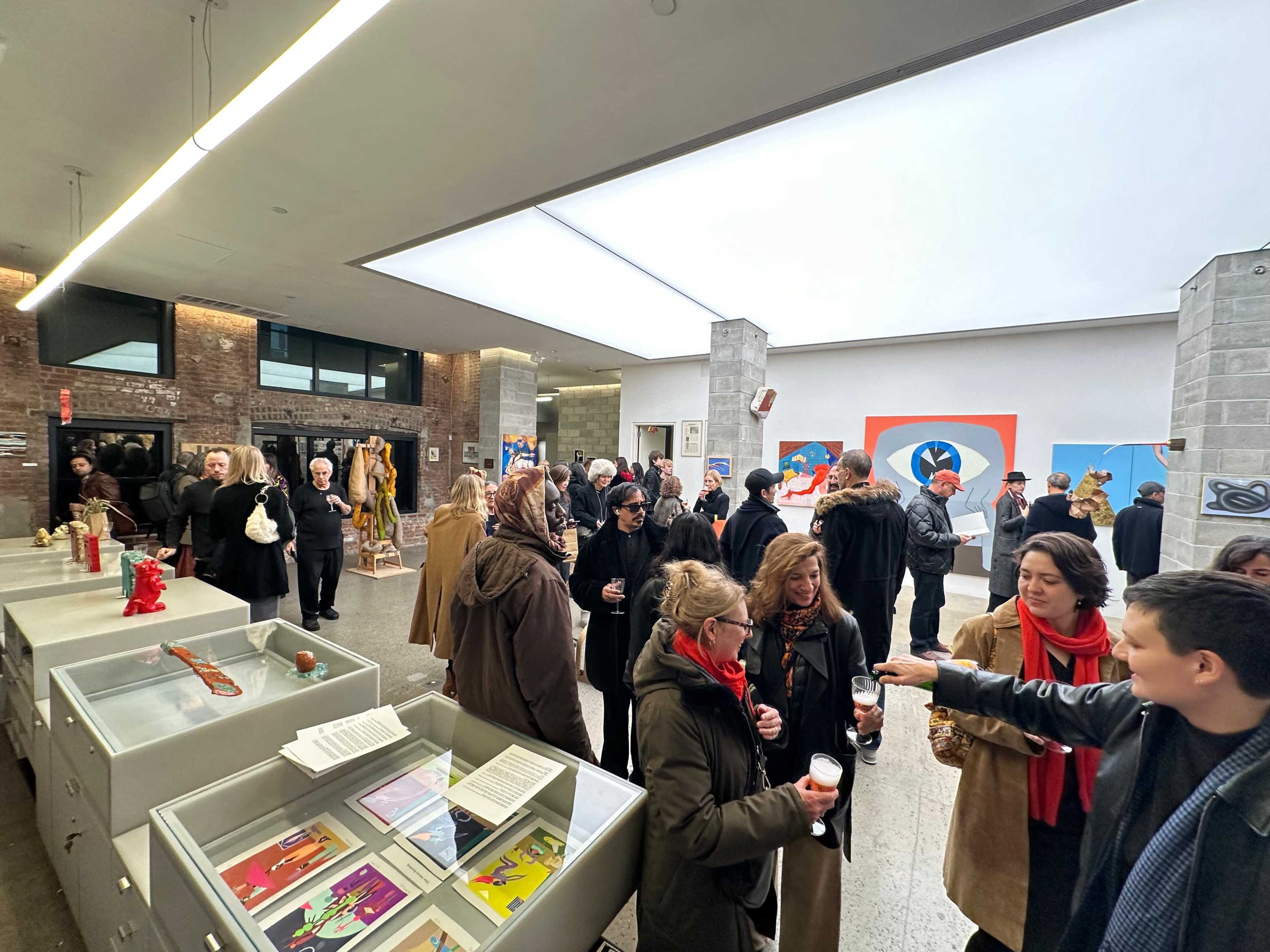 A diverse crowd gathers in an art gallery, engaged in conversation and viewing various artworks displayed on the walls and in glass cases.