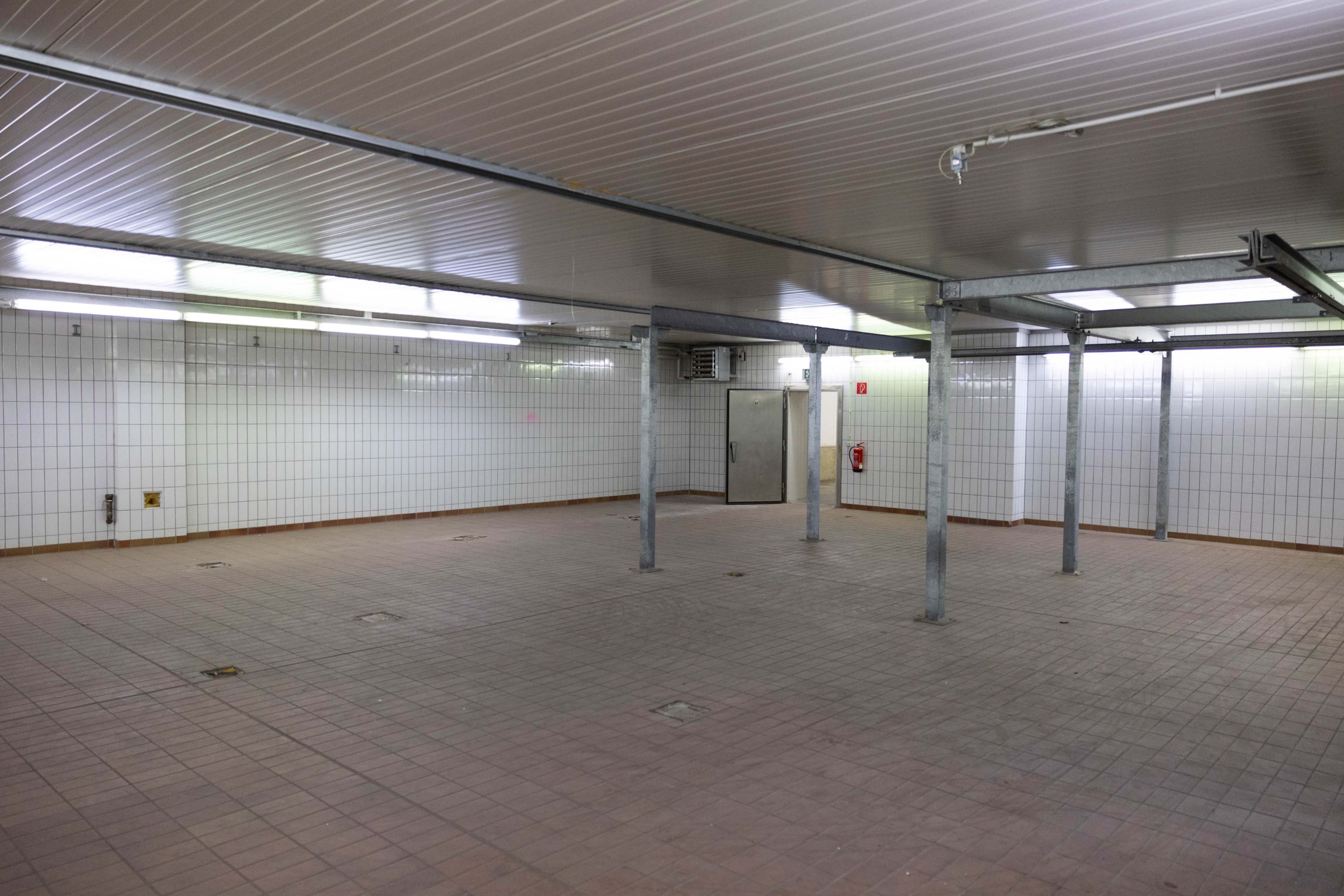 The image shows an empty, tiled indoor space with metal support columns and fluorescent lighting.