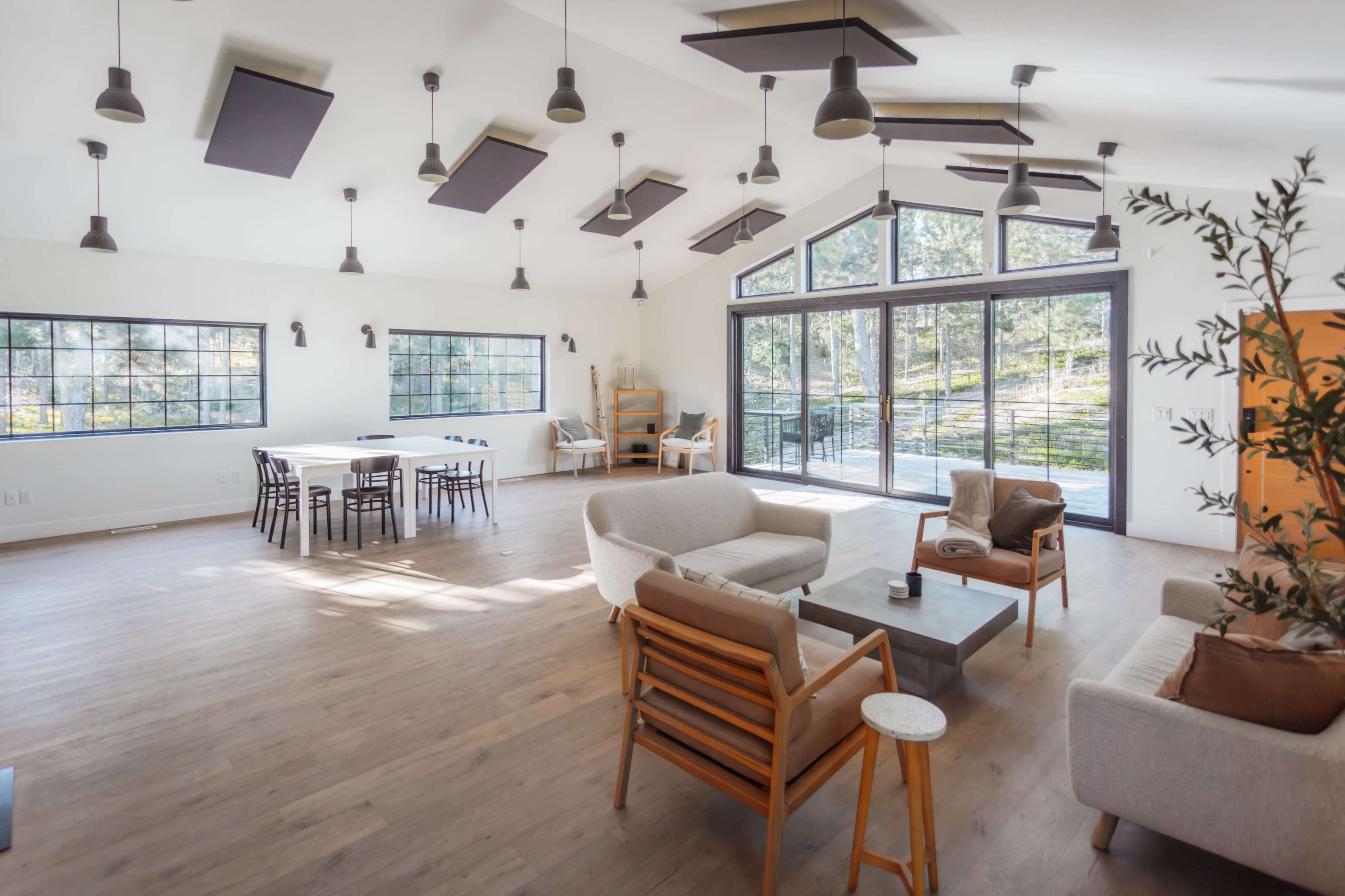 A spacious, well-lit living area features modern furniture, large windows, and an open layout with a dining table and access to an outdoor view.