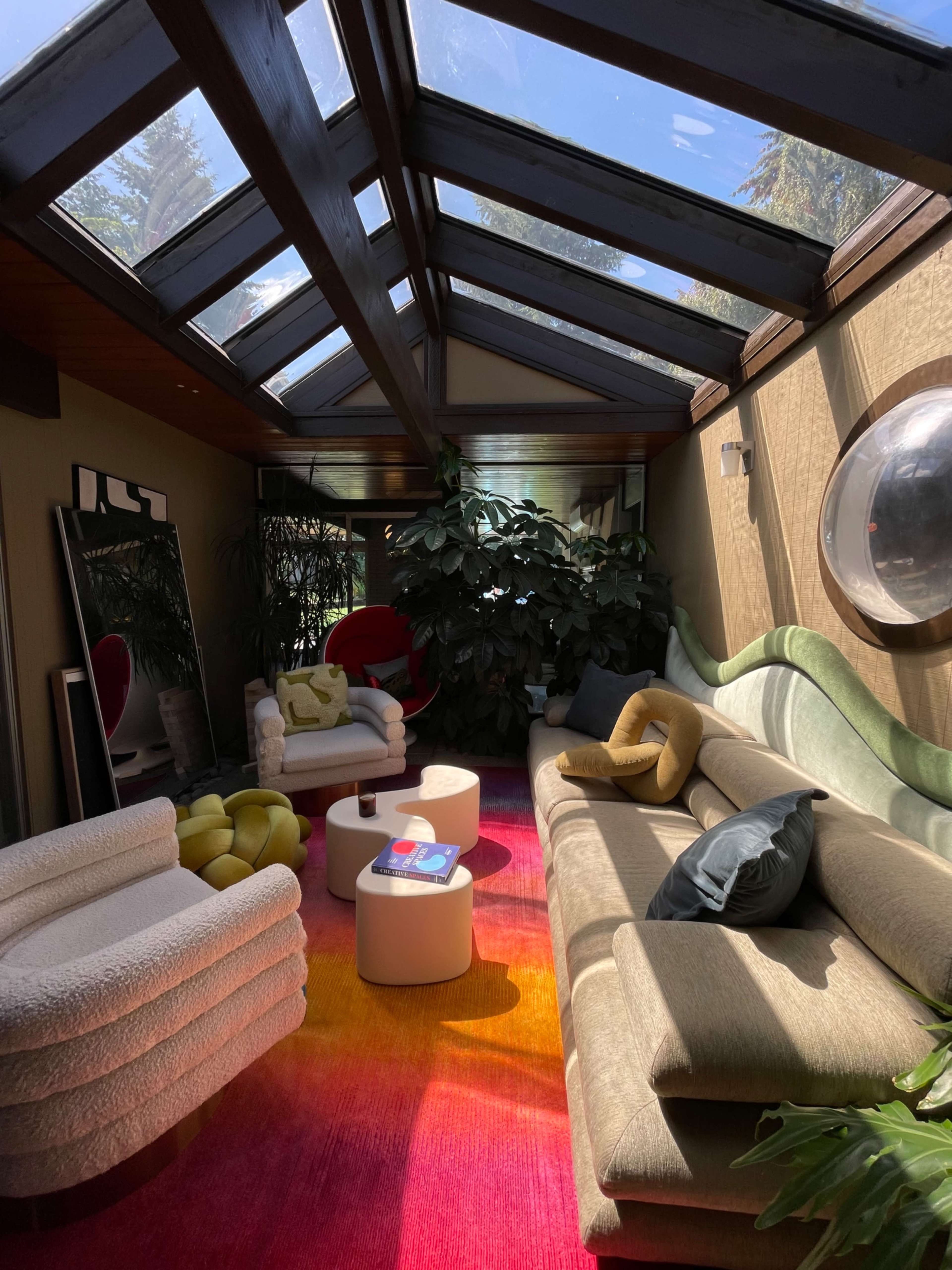 The scene features a sunlit room with a glass roof, containing modern seating arrangements, a vibrant rug, and several indoor plants.