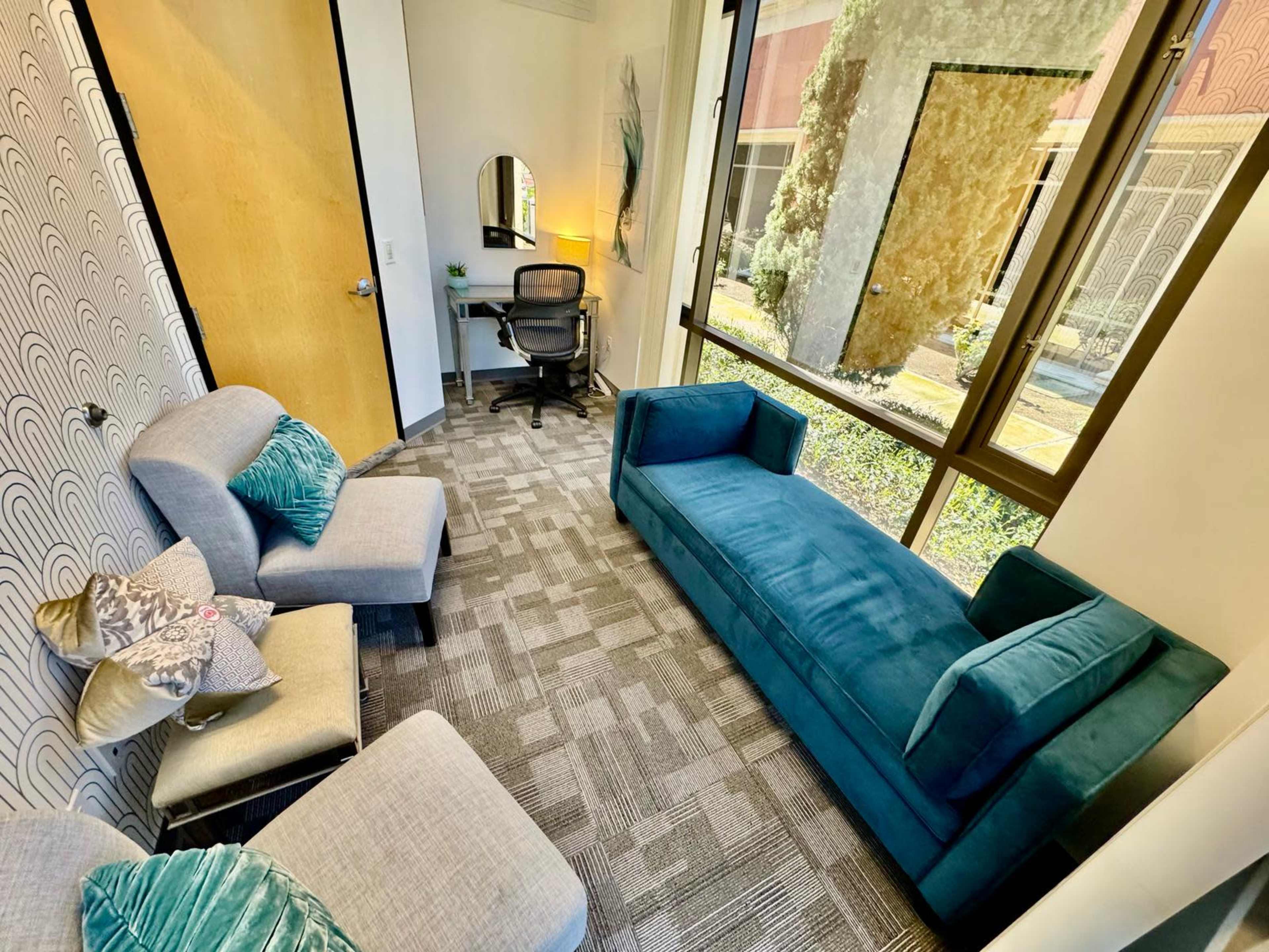 The image shows a small, well-lit office space featuring a blue couch, two grey chairs, and a desk with a chair, surrounded by large windows.