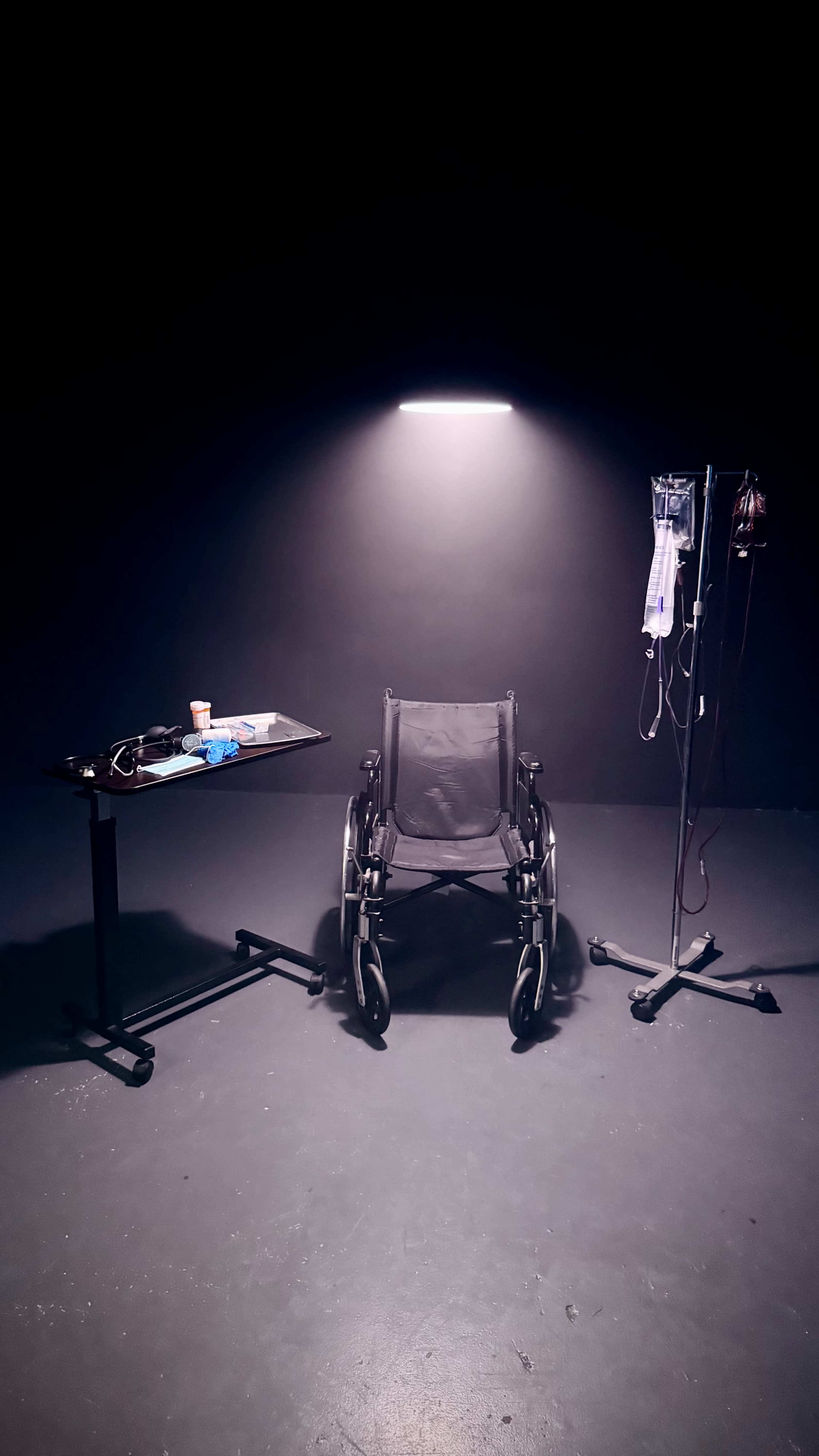 A wheelchair is positioned between a medical stand with IV bags and a small table with medical supplies in a dimly lit room.