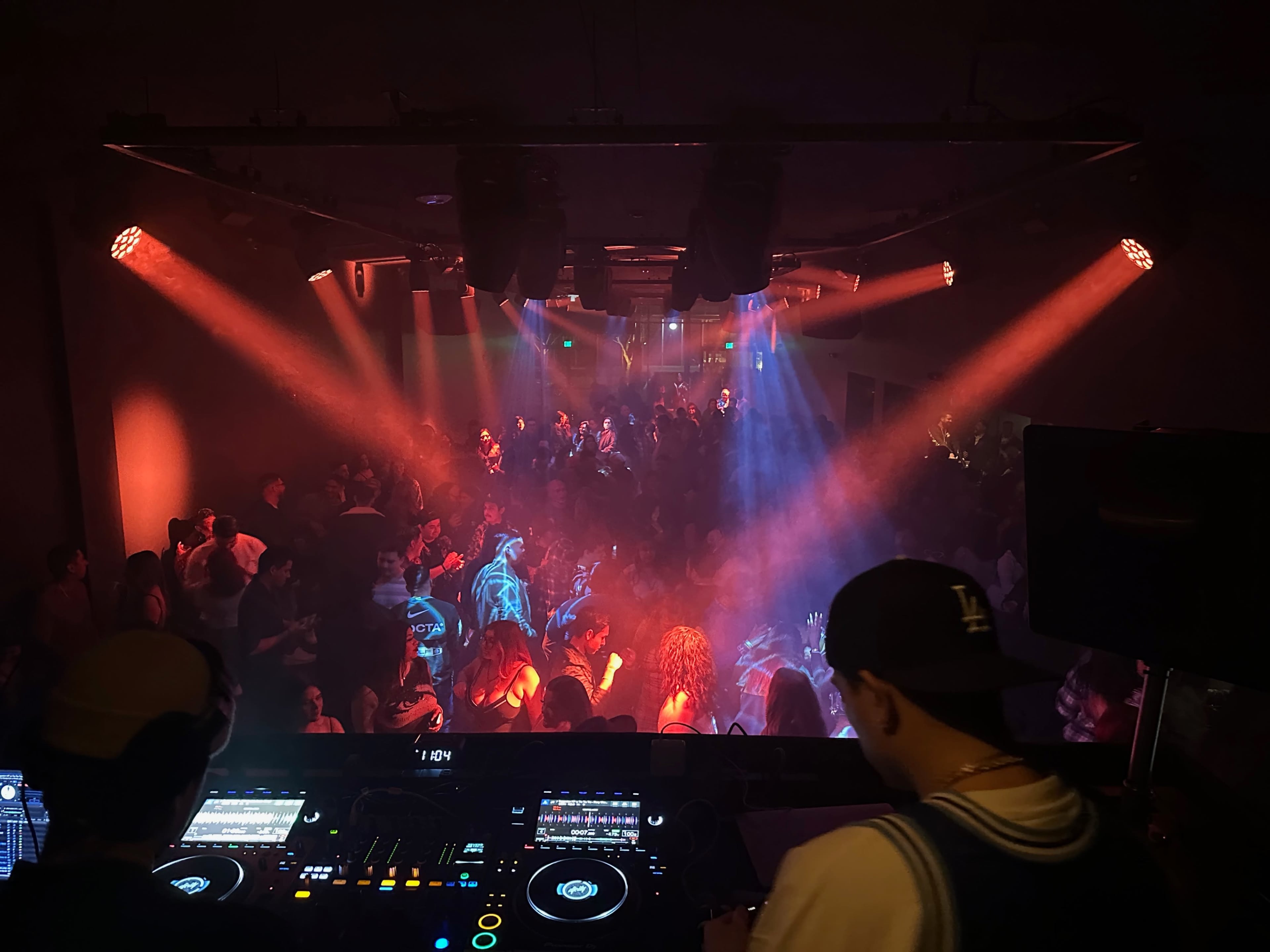 A DJ performs in a club while a crowd dances under colorful lights and fog.