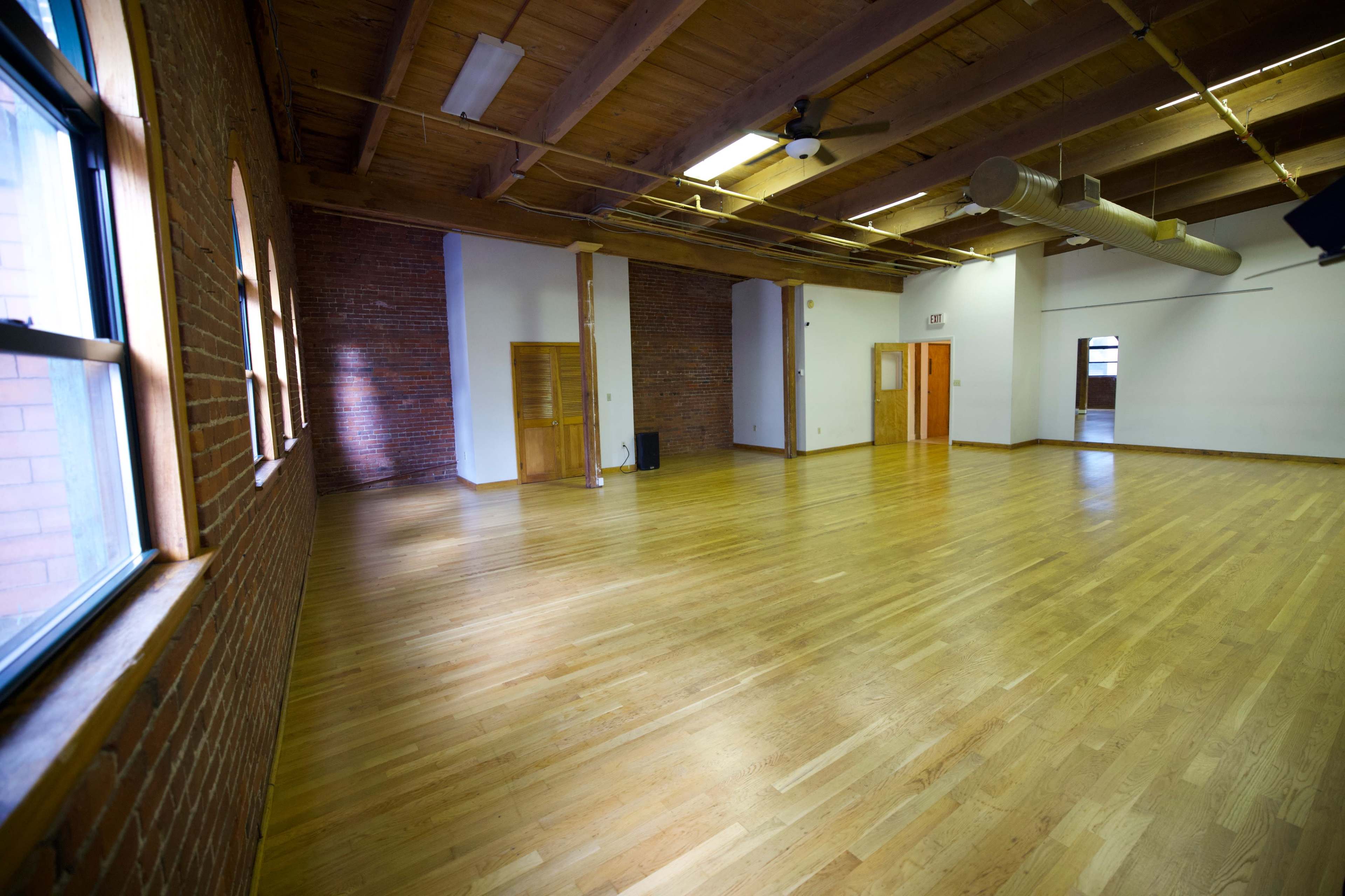 The image shows a spacious, empty wooden-floored room with exposed brick walls and large windows.