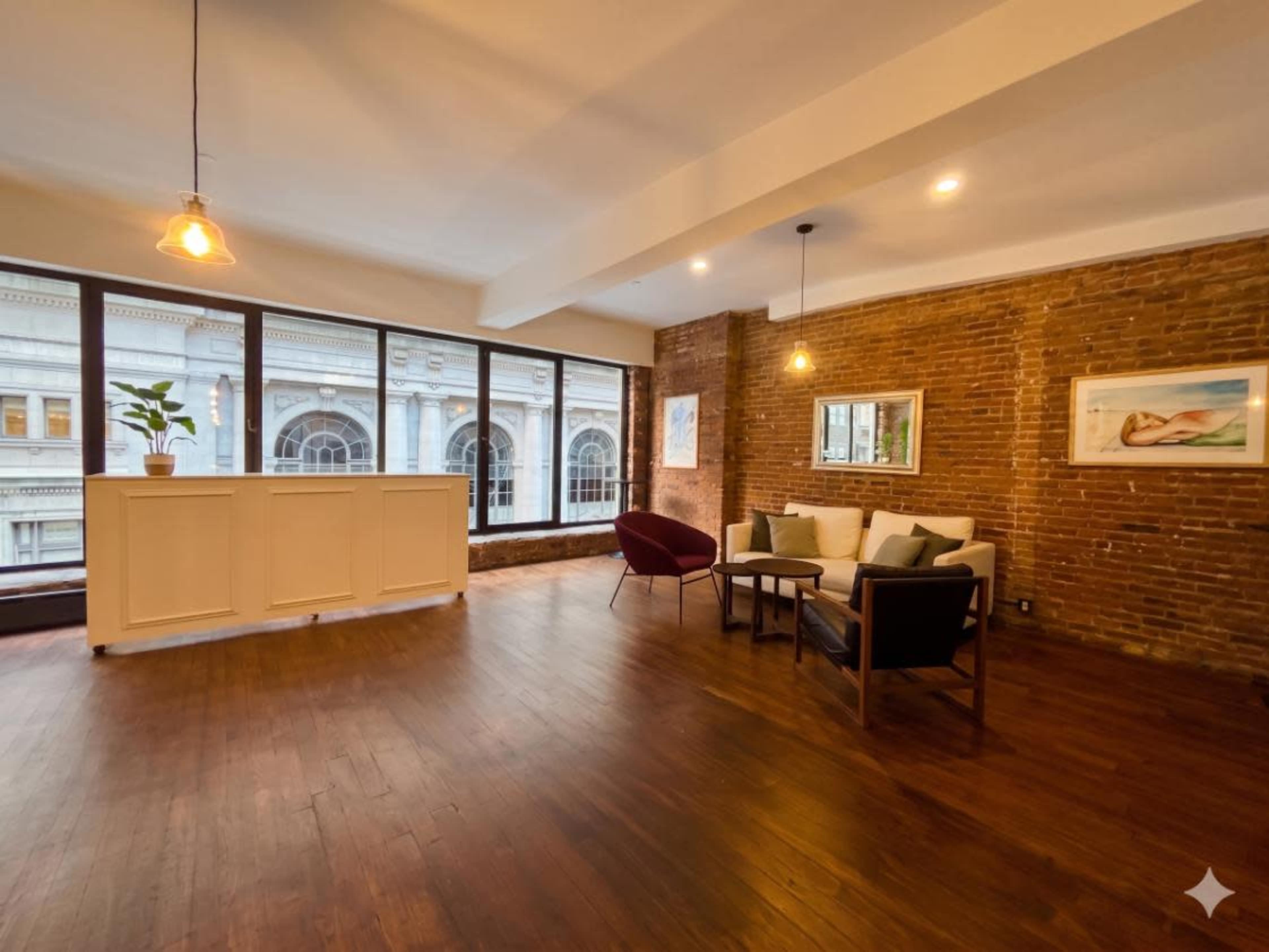 The image shows a spacious, modern living area with exposed brick walls, large windows, and minimalist furniture.