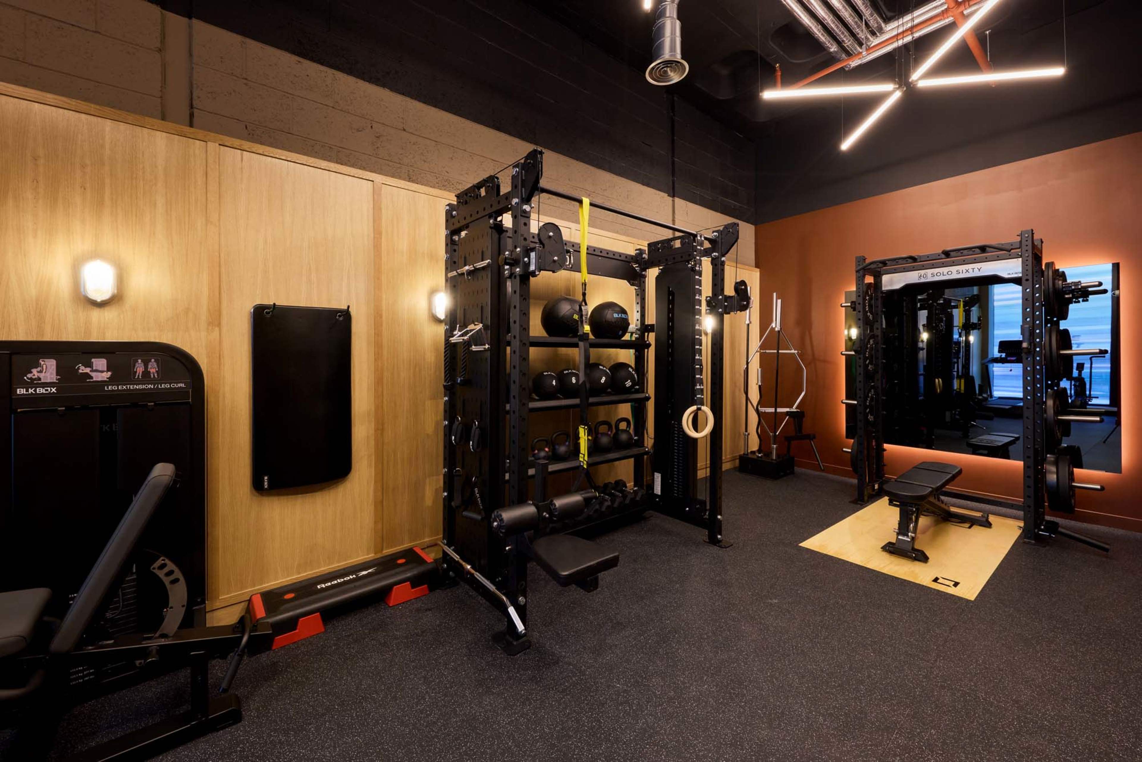 The image shows a well-equipped gym space featuring a multi-functional weight rack, various exercise machines, and free weights against a backdrop of wooden and dark walls.