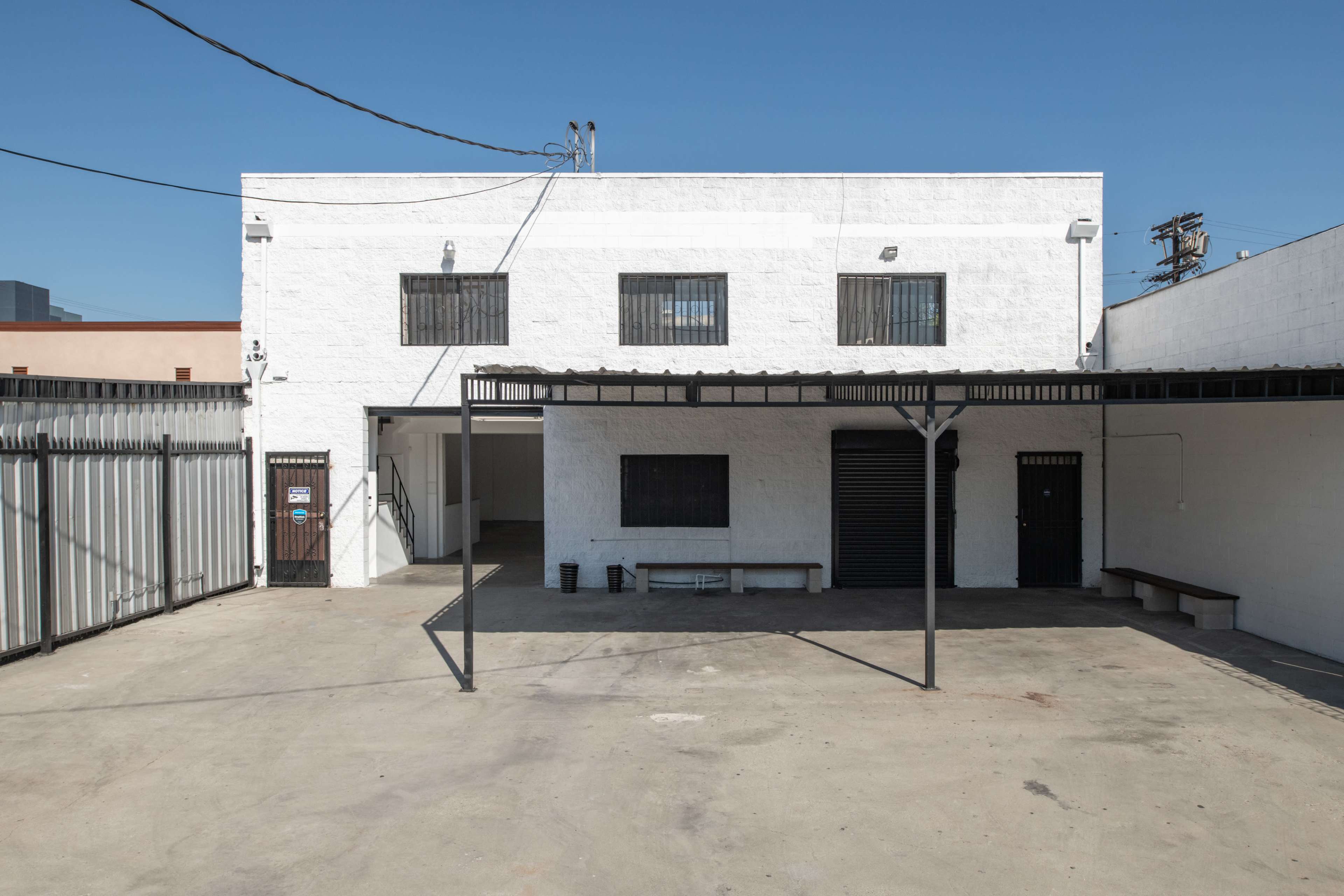 A spacious, vacant outdoor area in front of a white building with barred windows and a roofed structure supported by metal beams.