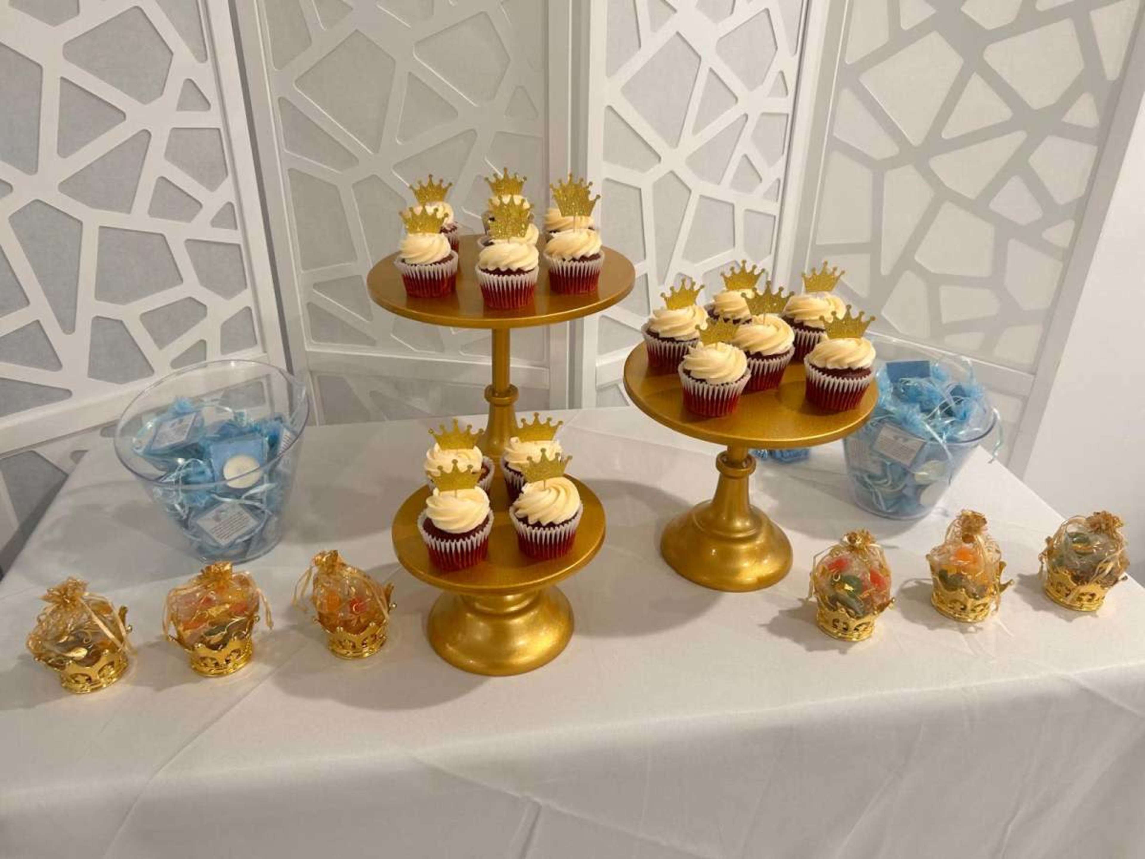 The image features a dessert display with two golden tiered stands holding red velvet cupcakes topped with cream cheese frosting, along with small decorative crowns, and two bowls containing blue candy.