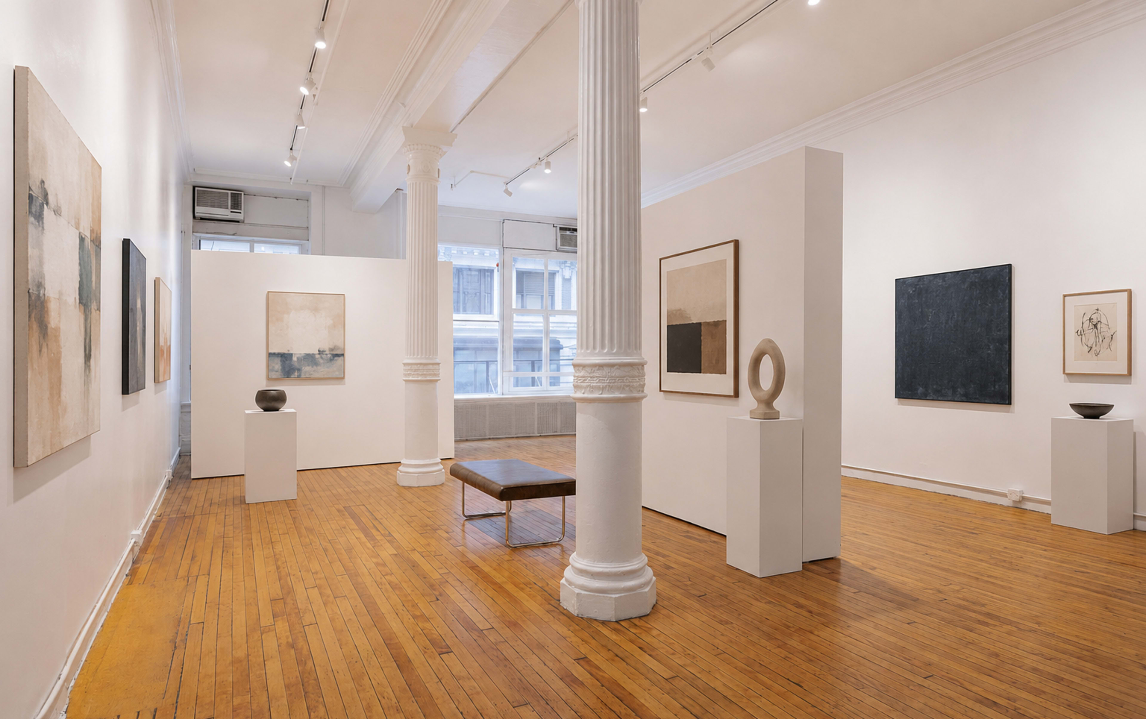 A spacious art gallery features abstract paintings on white walls, with sculptures displayed on pedestals and a wooden bench in the center.
