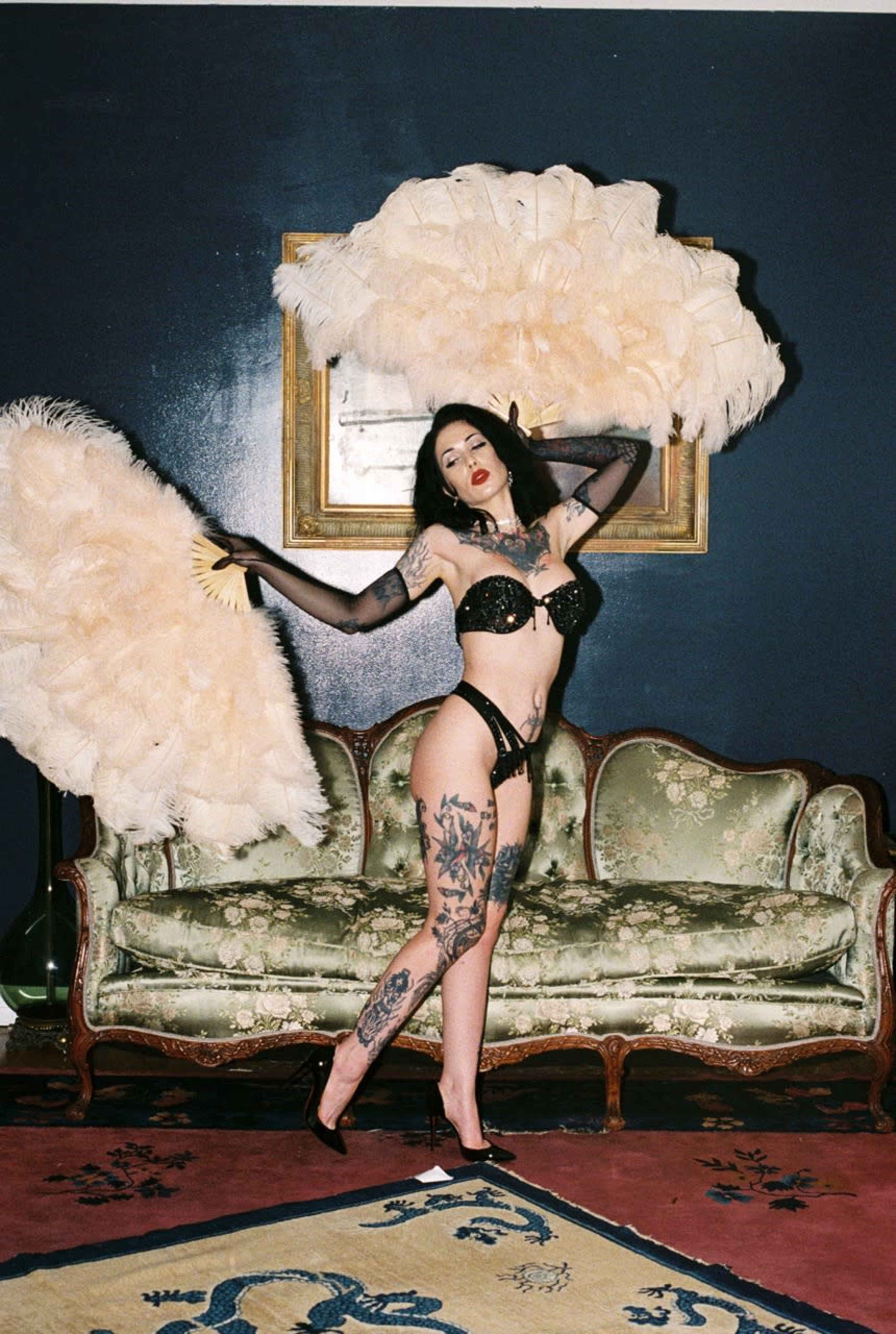 A woman poses with two large feather fans while standing in front of a vintage couch in a dimly lit room.