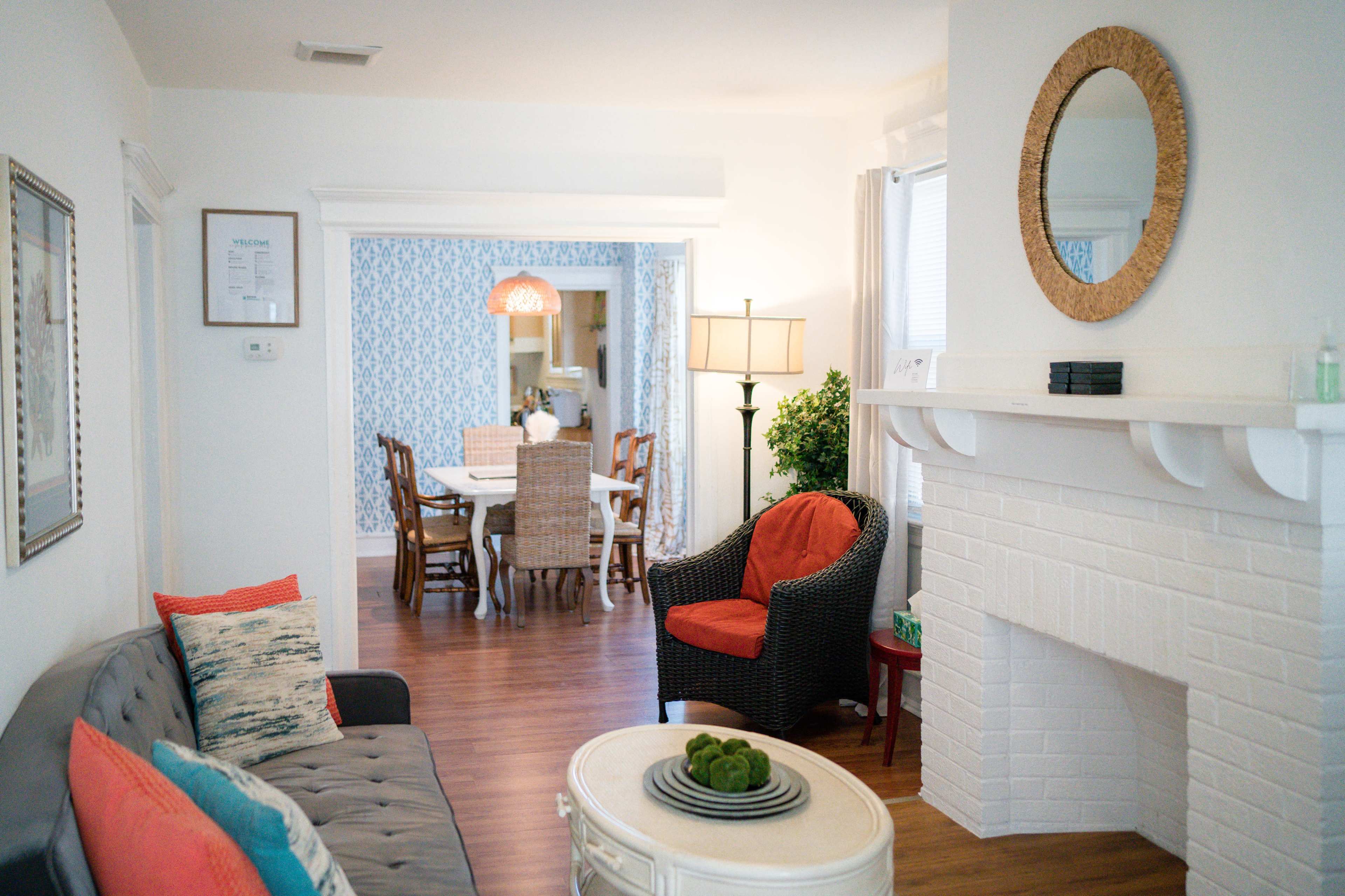 A bright living room with a gray couch, a round white coffee table, and a wicker chair, leading to a dining area with a table set for six.