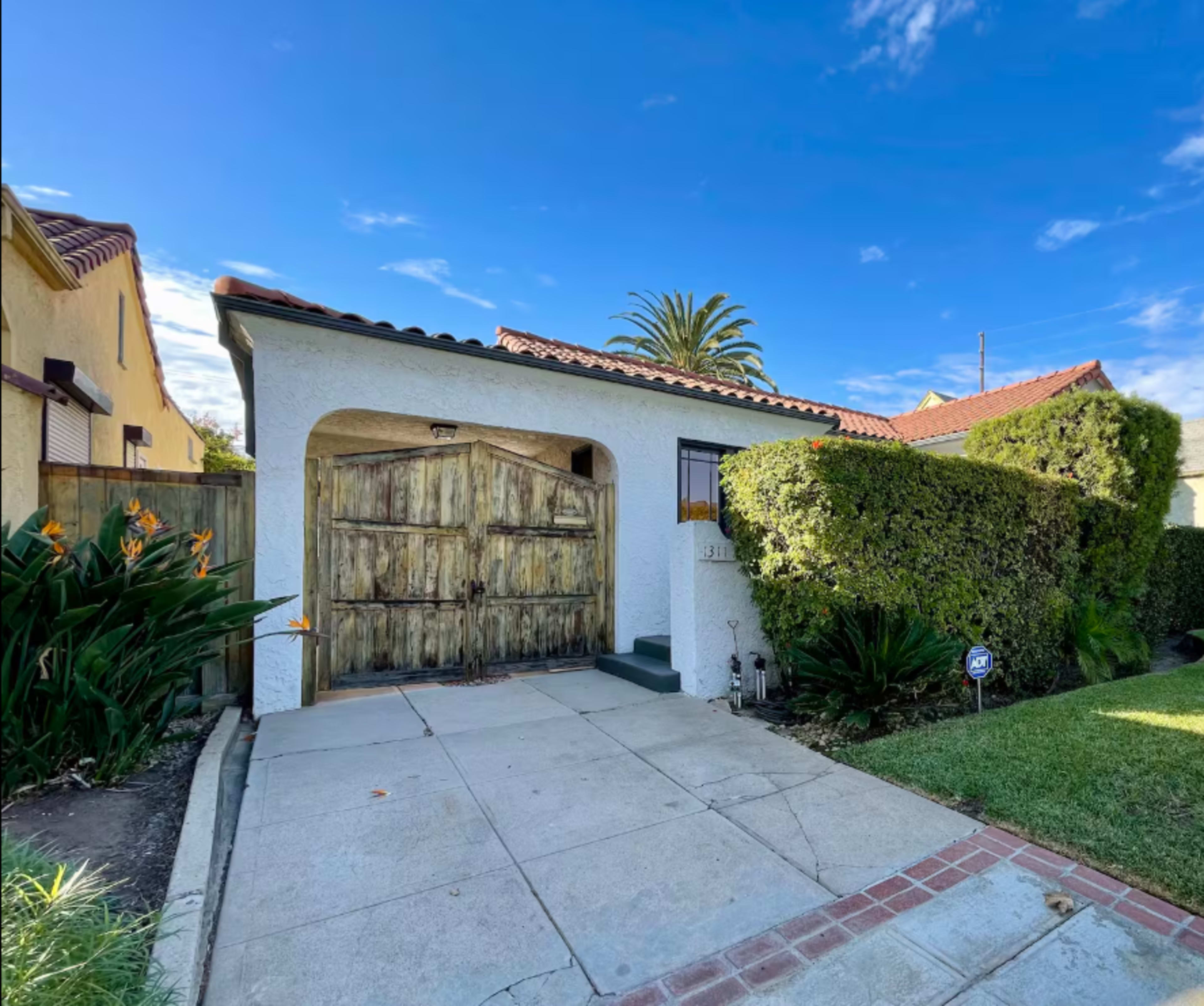 A single-story house with a wooden front door, surrounded by manicured hedges and palm trees, sits on a paved driveway under a blue sky.