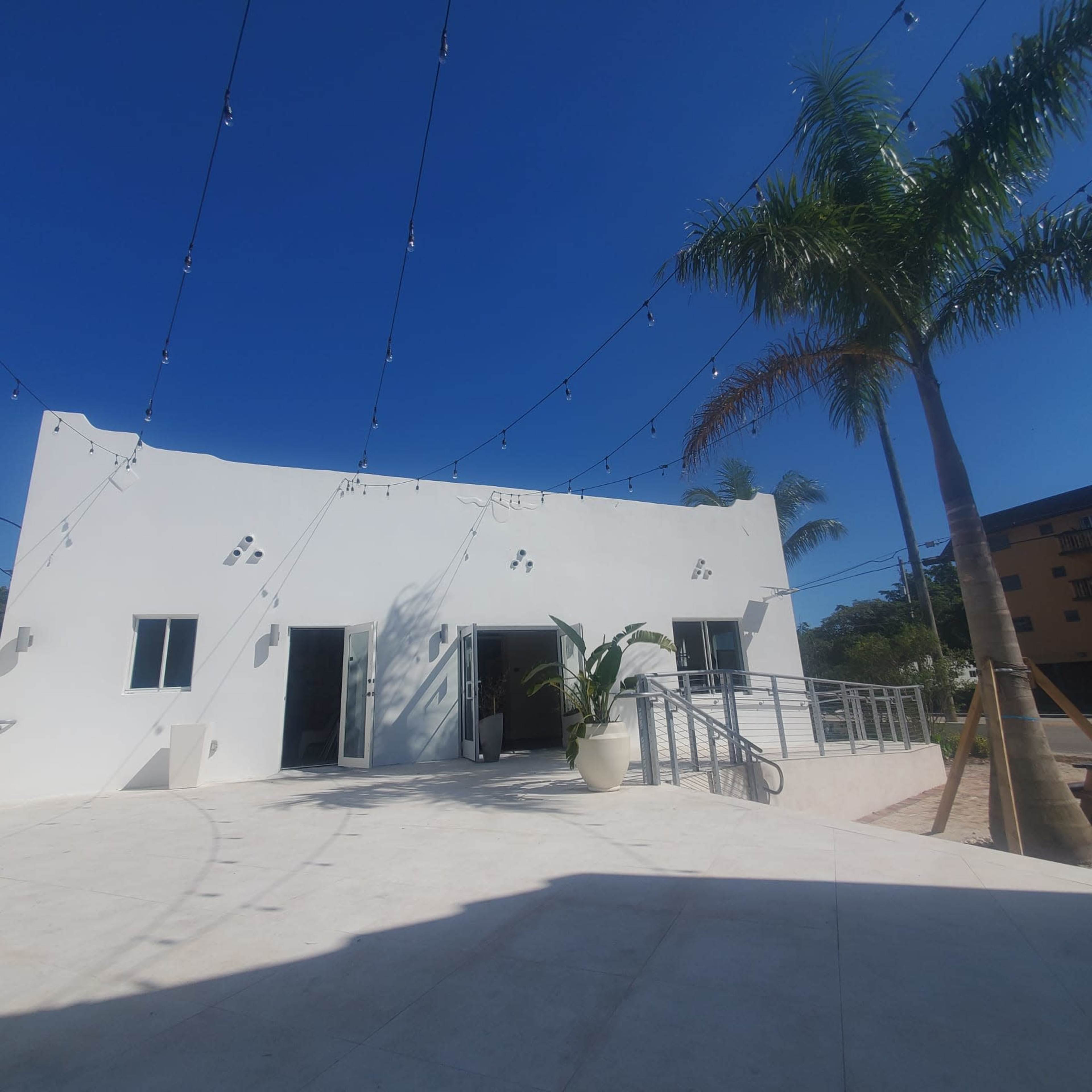 A modern white building with large windows and a patio is shown under clear blue skies, surrounded by a palm tree and string lights overhead.