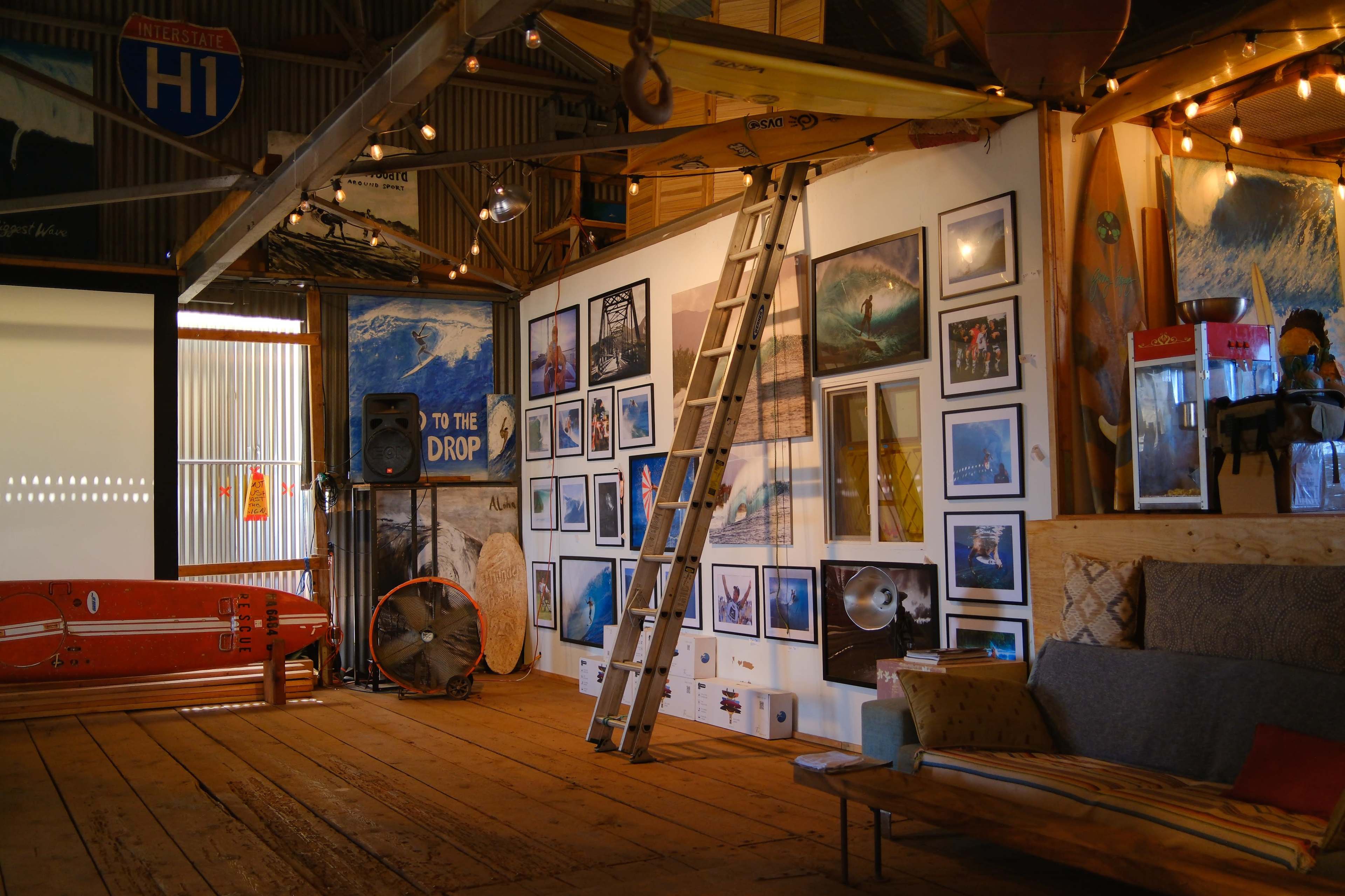 The interior of a rustic building features a vertical ladder leaning against a wall adorned with various framed photographs, with surfboards and a fan visible in the space.