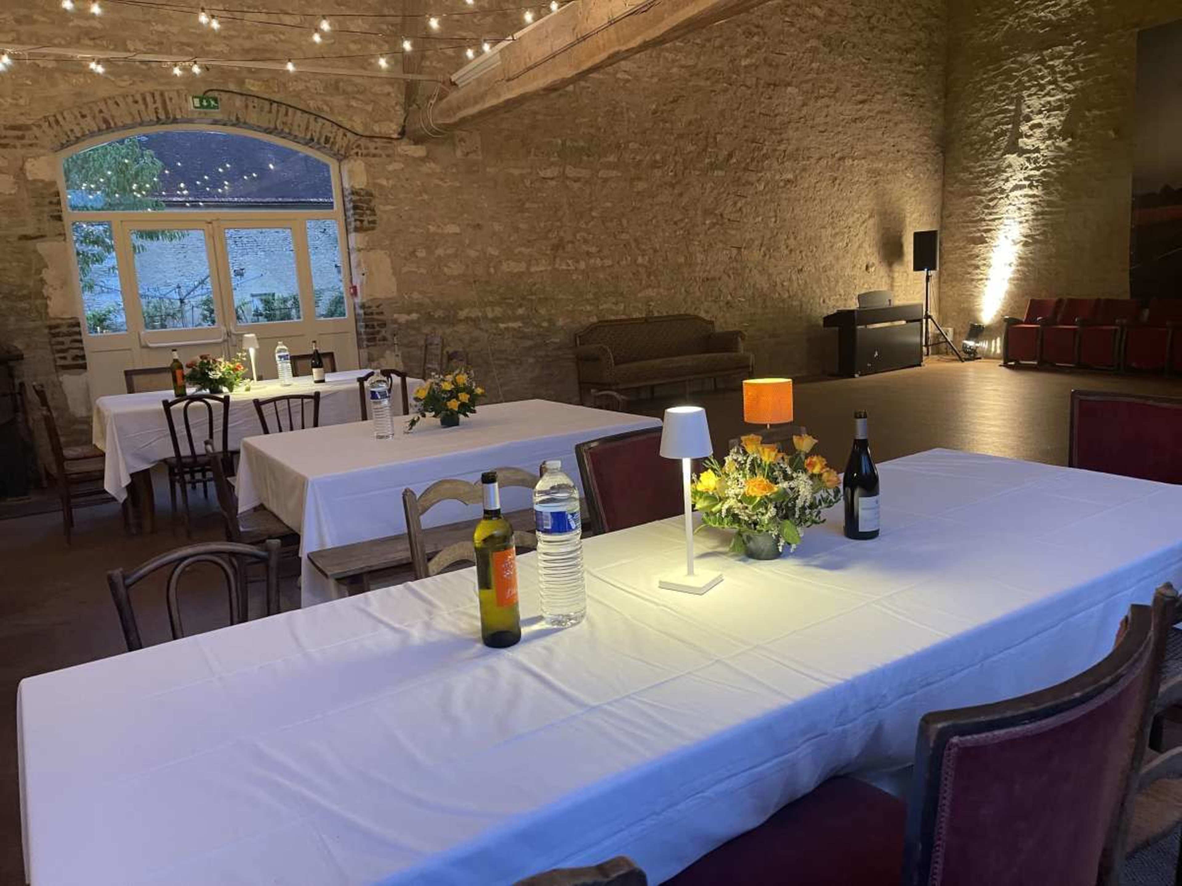 The image shows a spacious event room with several tables covered in white tablecloths, adorned with flowers, and bottles of wine and water on each table.
