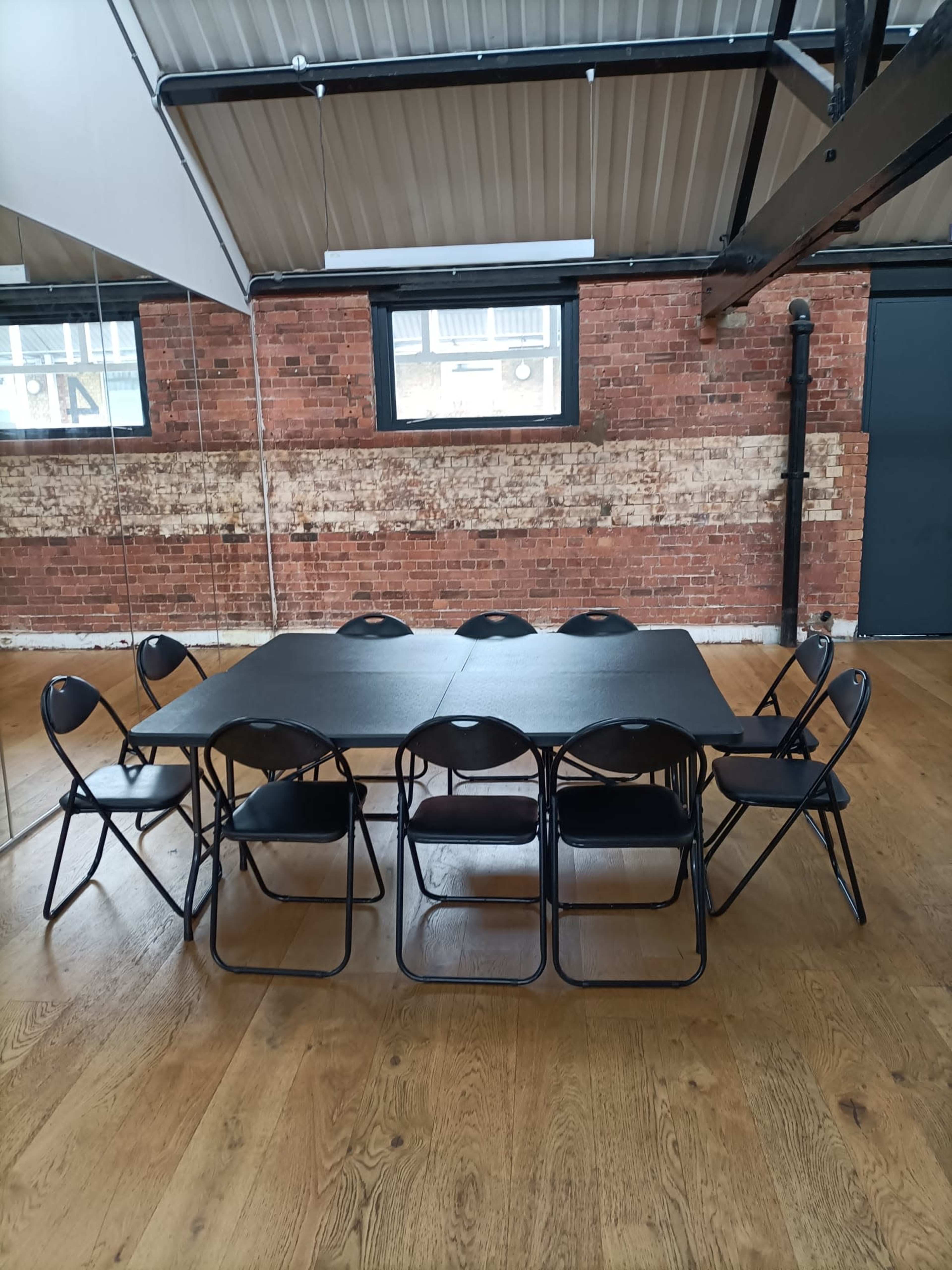 A rectangular table surrounded by eight black folding chairs is positioned in a room with brick walls and wooden flooring.