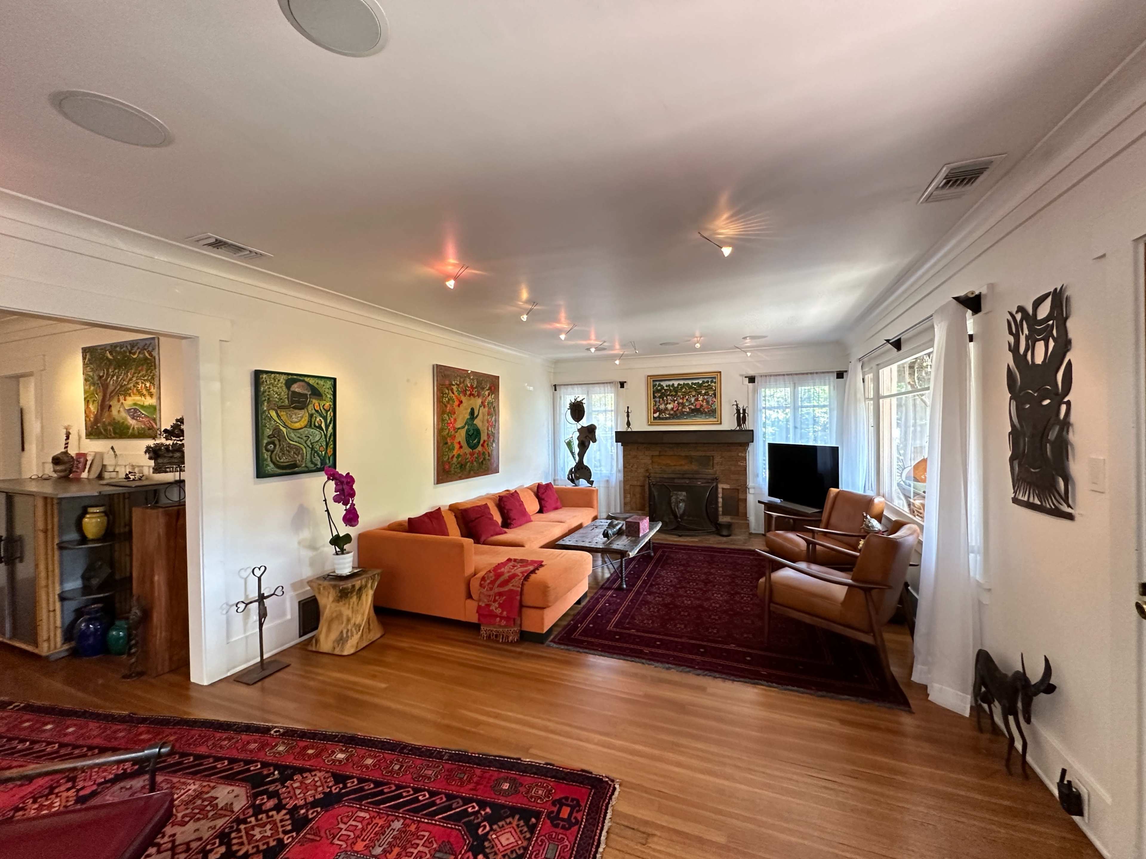 The image shows a spacious living room featuring an orange sectional sofa, wooden furniture, colorful artwork on the walls, and a fireplace.