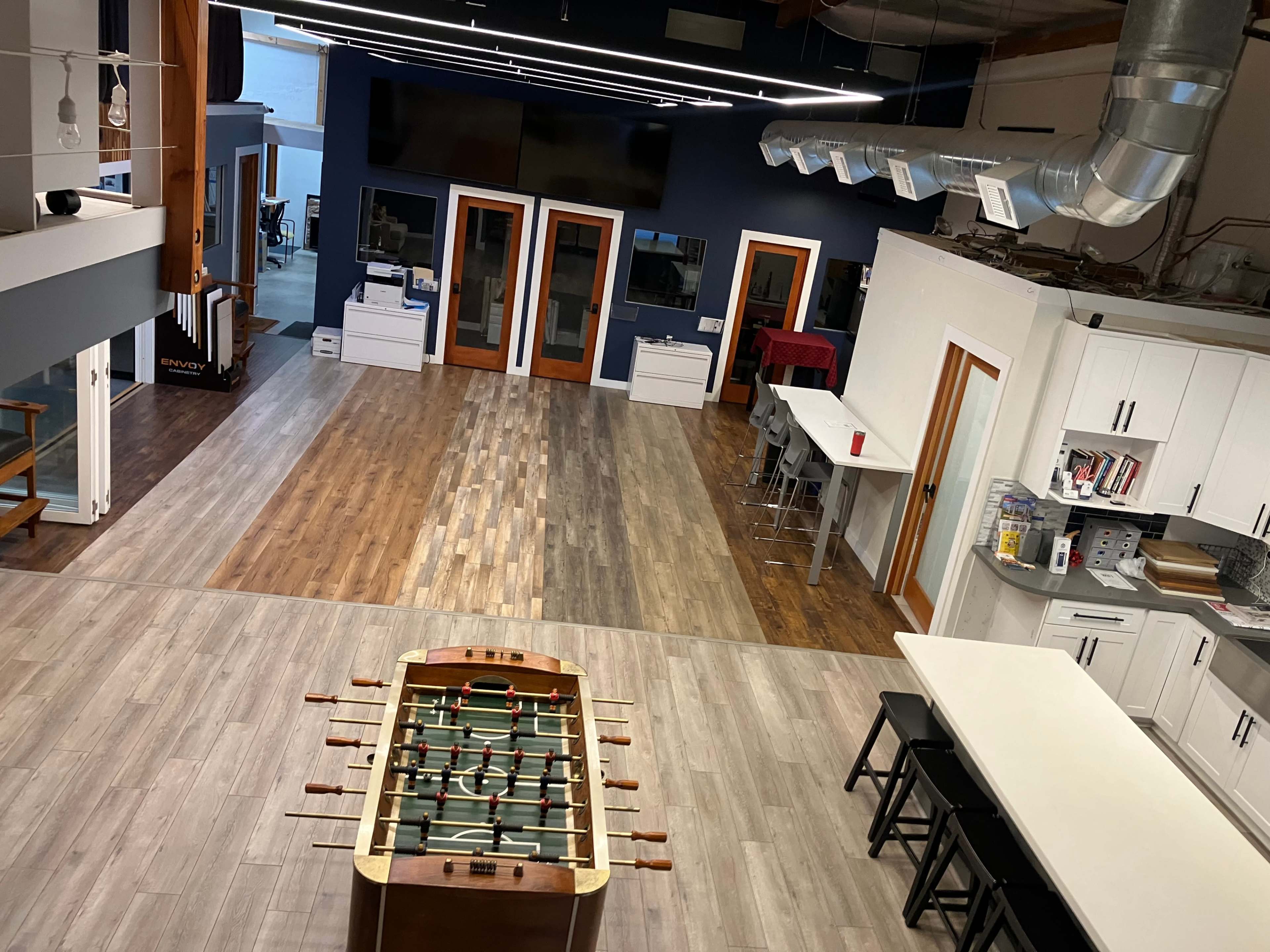 The image shows a large, open office space featuring a foosball table, wooden flooring in various tones, and modern kitchen amenities.