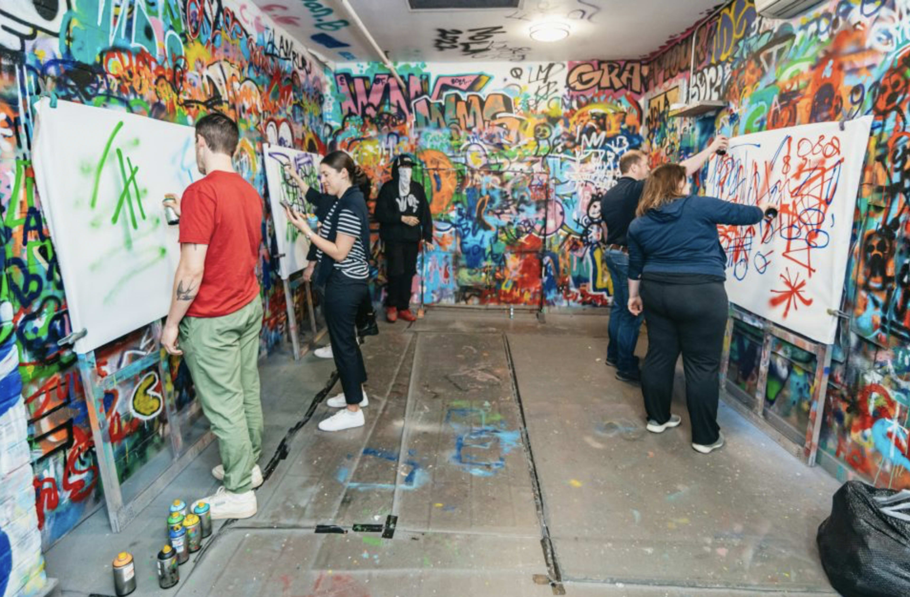 A group of people is engaged in painting graffiti on large white canvases in a brightly colored, graffiti-covered room.